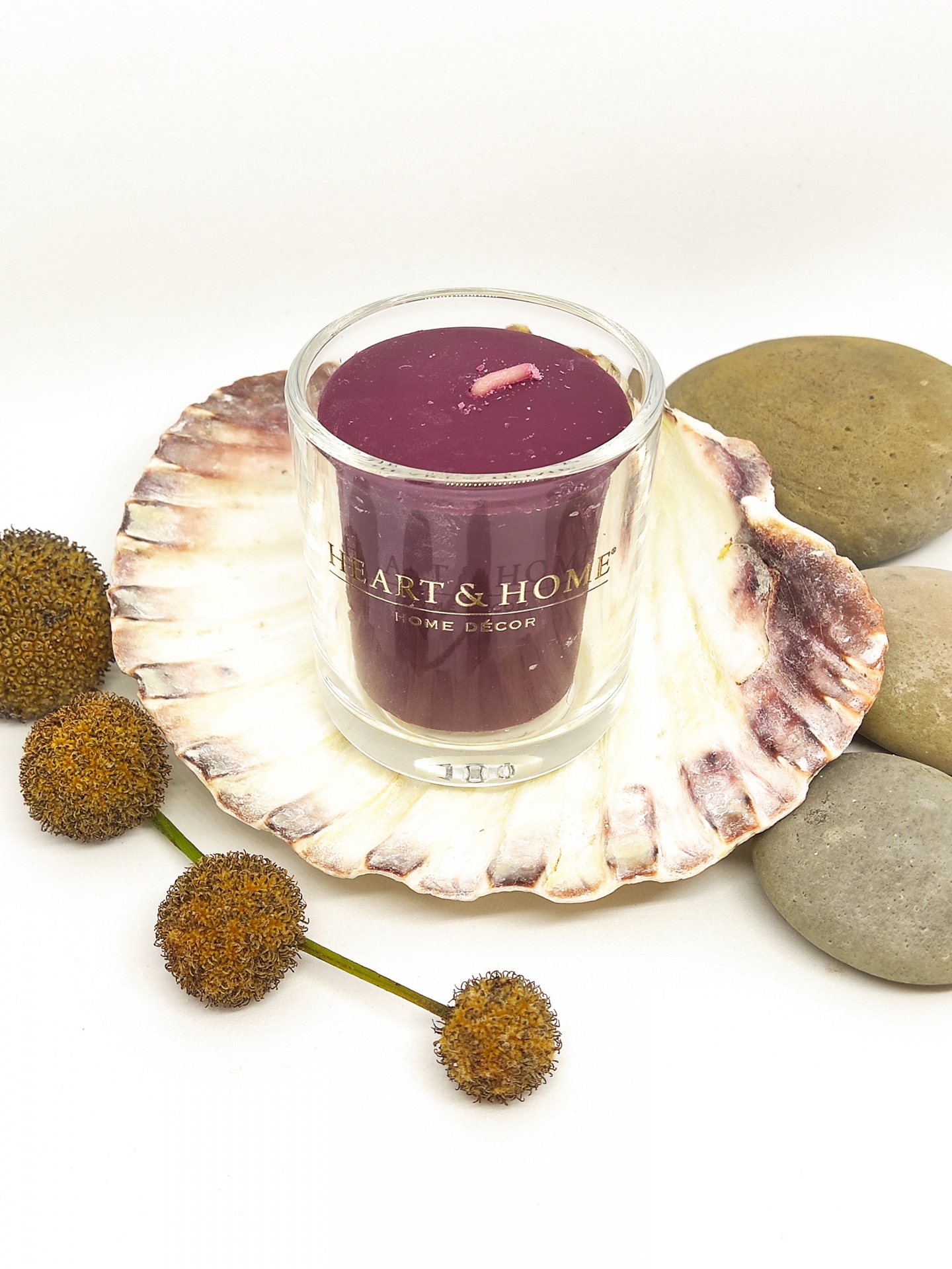 Vela roxa em copo de vidro com inscrição 'HEART & HOME' sobre concha com bolas secas e pedras