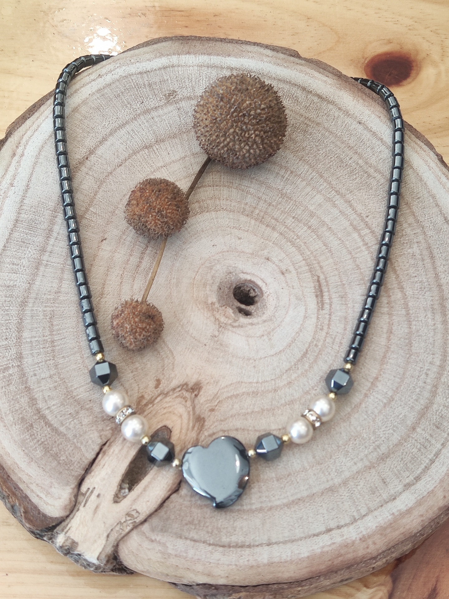 Colar com coração cinza metálico, contas prateadas e pérolas brancas sobre disco de madeira