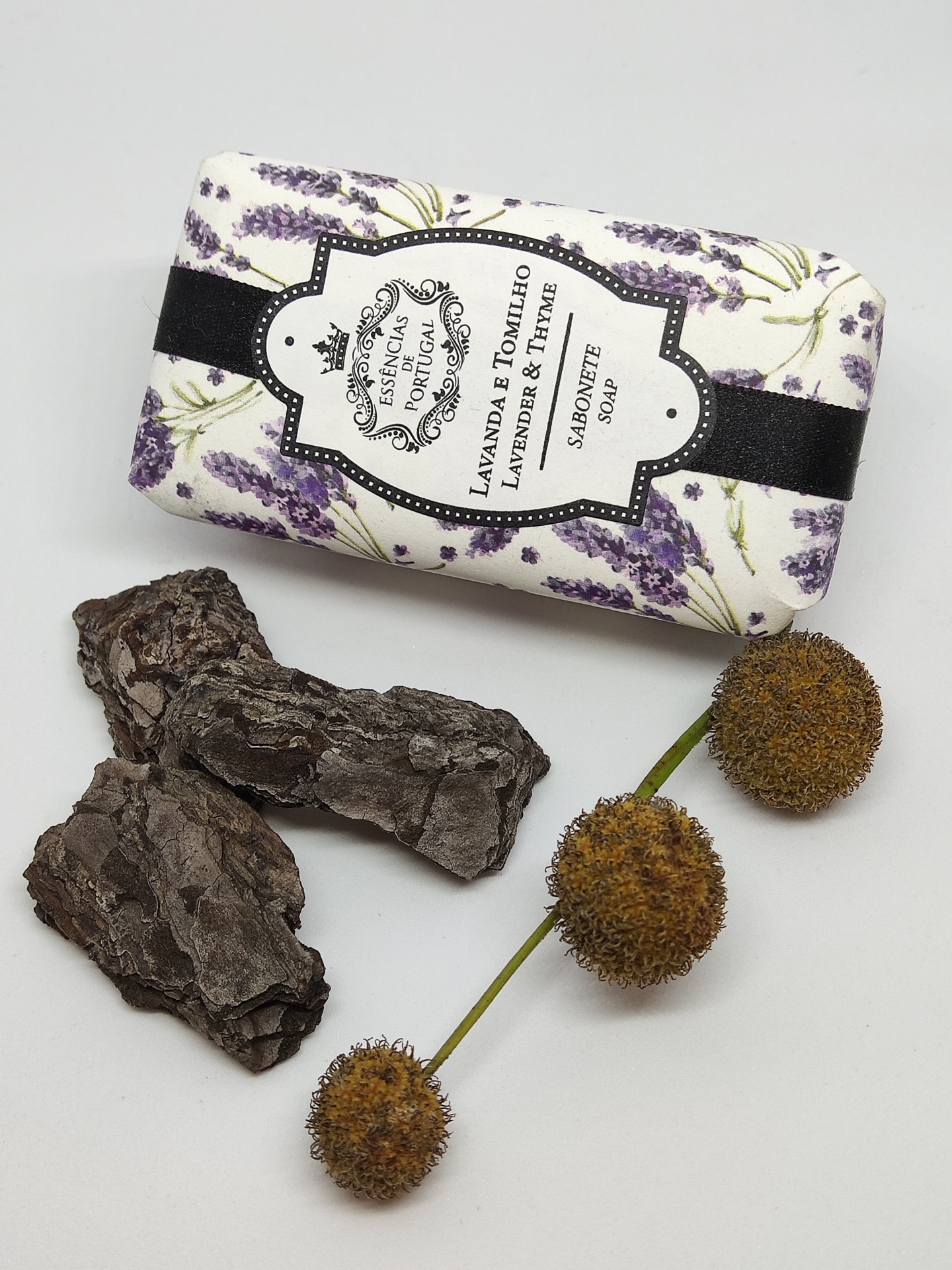 Sabonete com embalagem floral de lavanda, pedaços de casca de árvore escura e três esferas marrons secas em fundo branco
