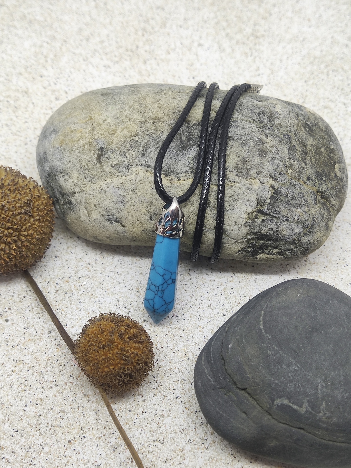 Colar com pendente de pedra azul turquesa e cordão preto sobre fundo de areia clara com pedras e elementos secos.