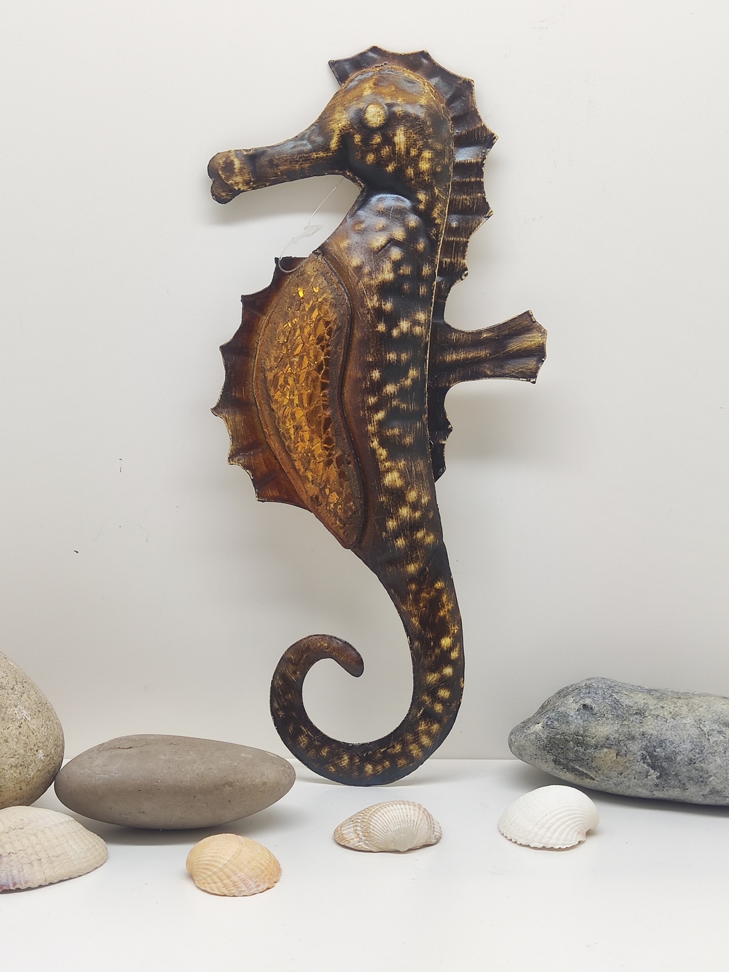 Escultura de cavalo-marinho em metal com pedras e conchas em fundo branco.