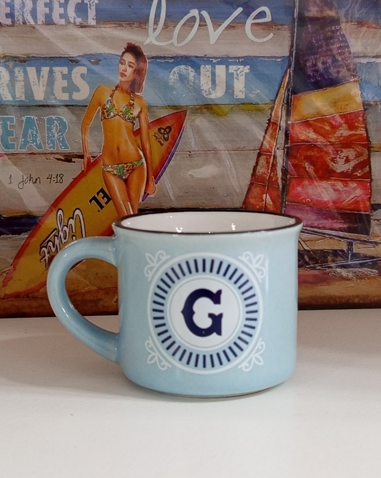 Caneca azul clara com letra G e fundo de uma imagem com mulher de biquíni e prancha de surf.