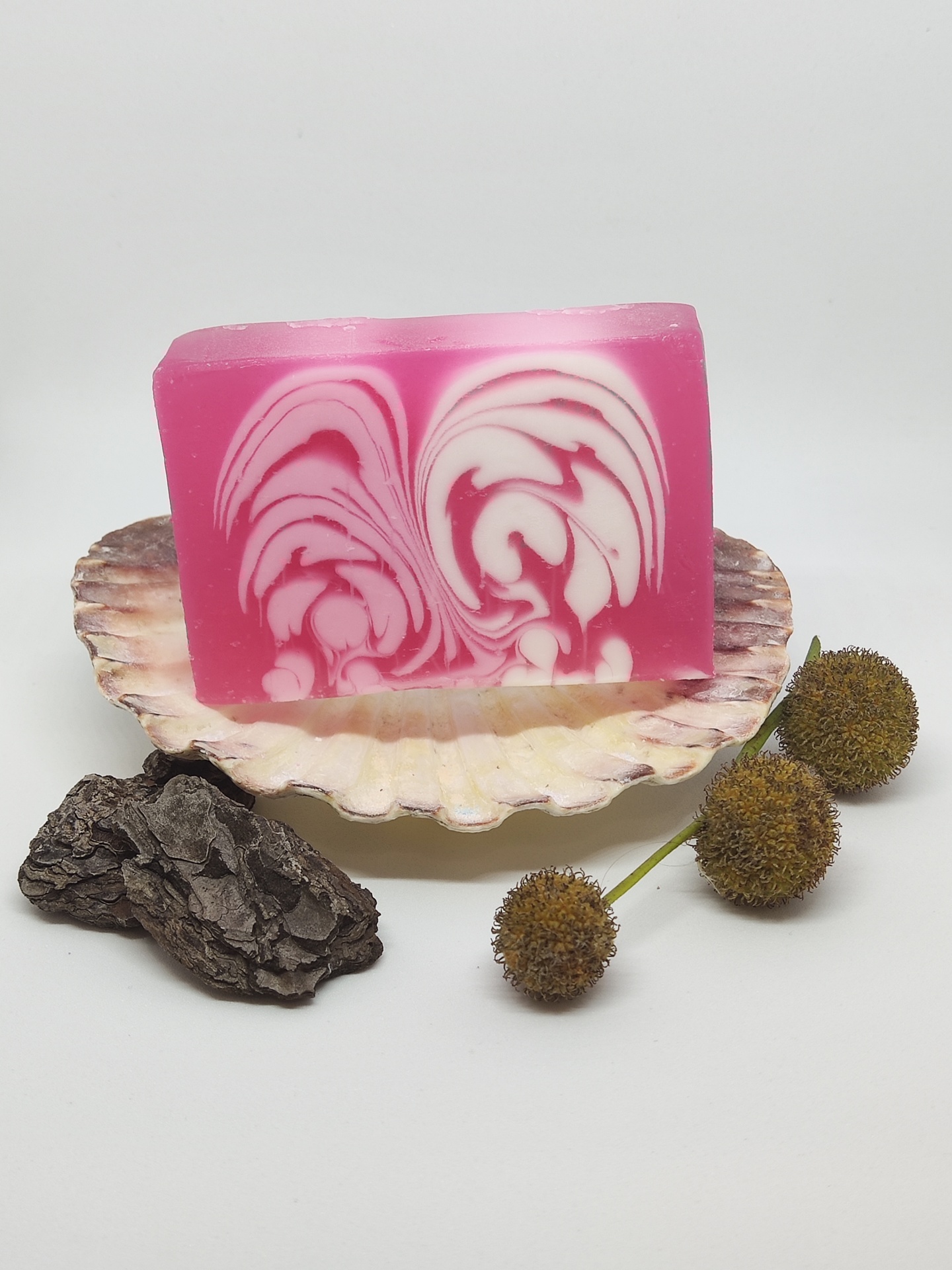 Sabonete marmoreado rosa e branco sobre concha com casca de árvore e flores secas.