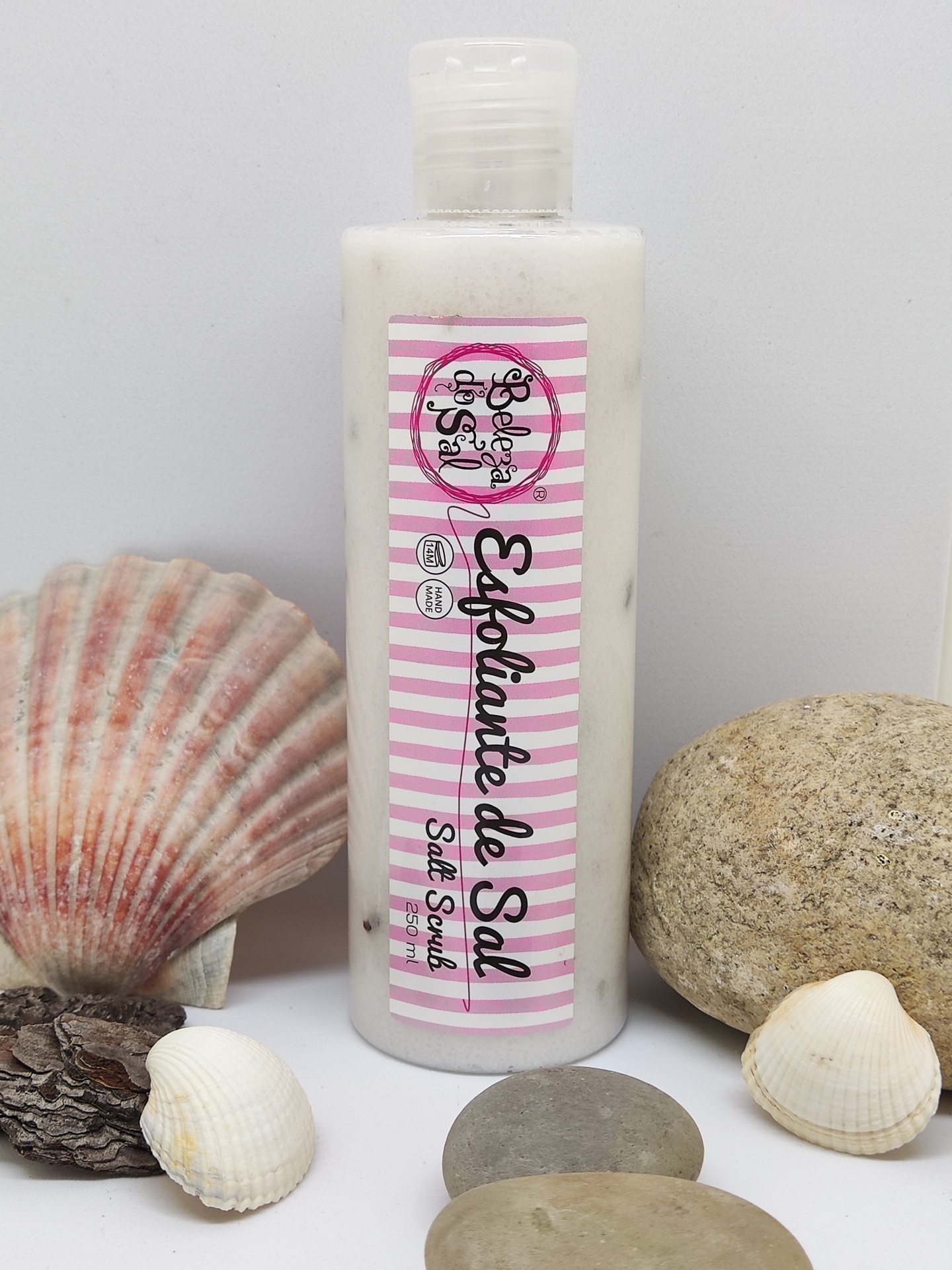 Frasco de esfoliante de sal com rótulo rosa e branco rodeado por conchas e pedras