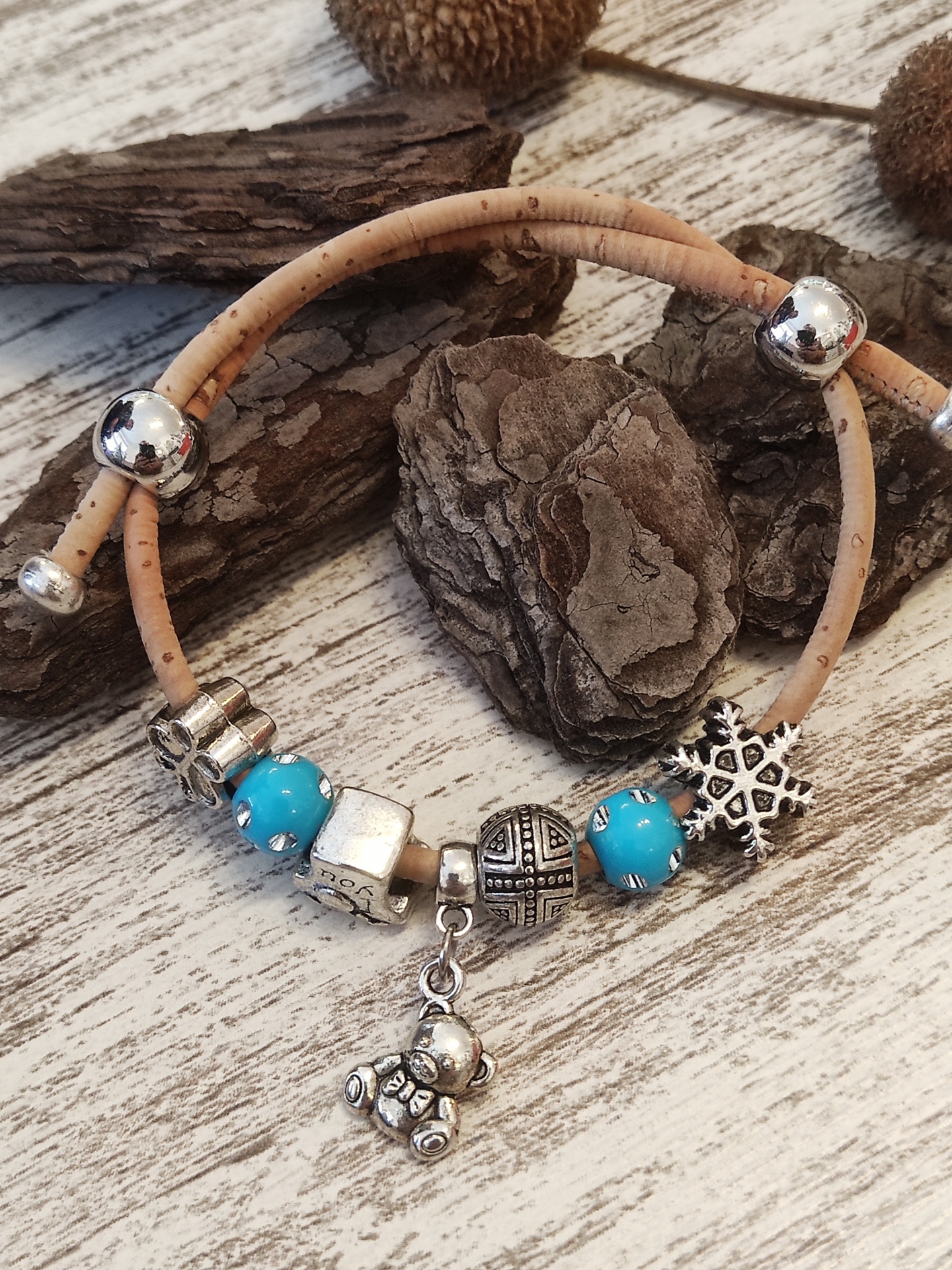 Pulseira de cortiça com contas prateadas e azuis sobre madeira clara e pedaços de casca