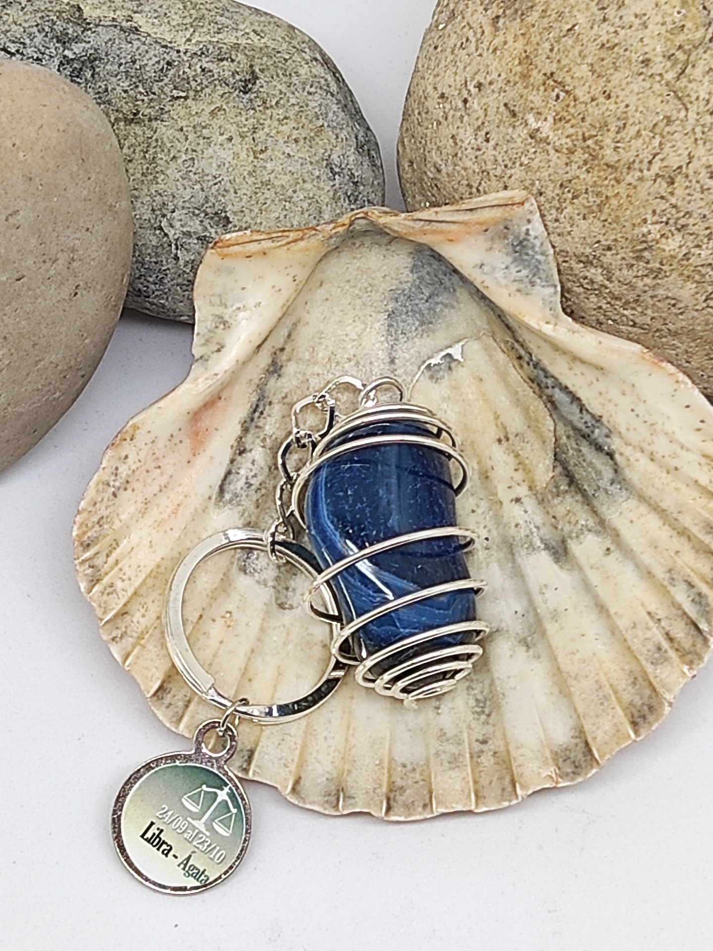 Chaveiro com pedra azul e pendente redondo sobre concha com pedras no fundo.