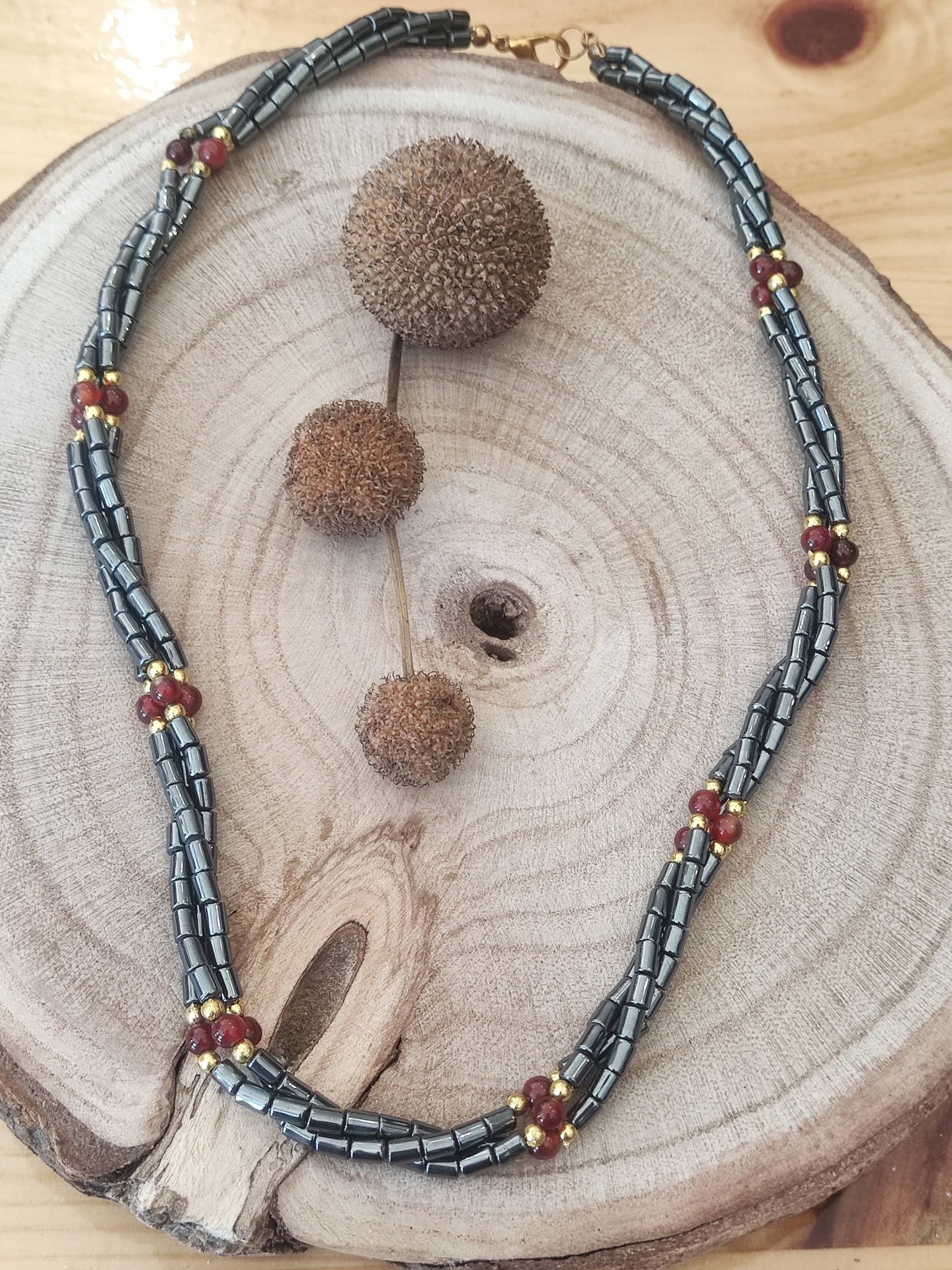 Colar com contas metálicas prateadas e esferas vermelhas e douradas sobre base de madeira clara