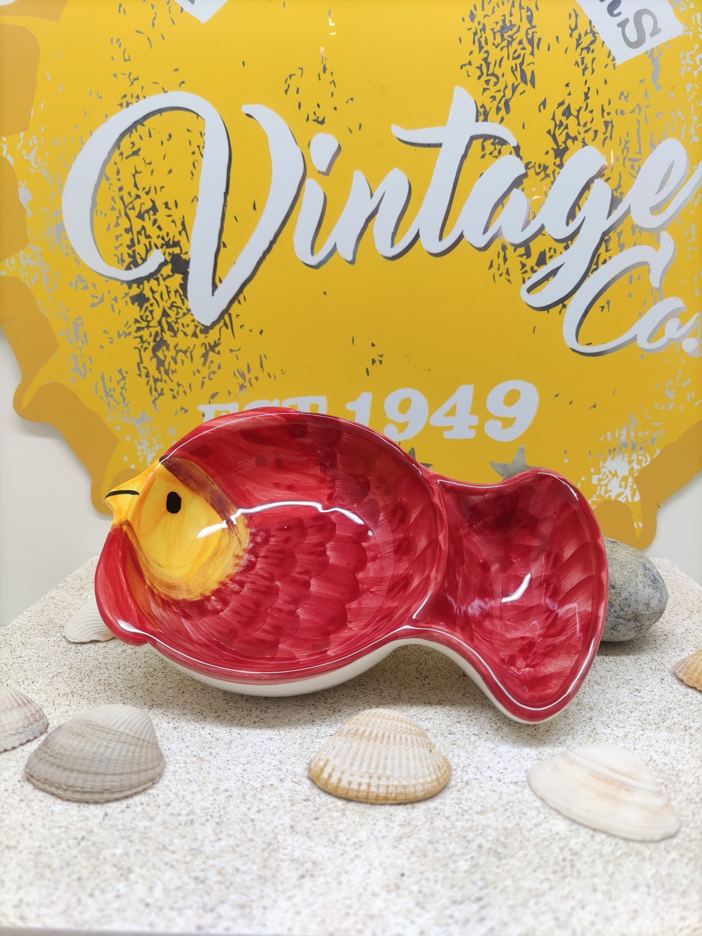 Prato cerâmico em forma de peixe vermelho com detalhes amarelos sobre areia e conchas, fundo amarelo com texto 'Vintage Co. EST. 1949'
