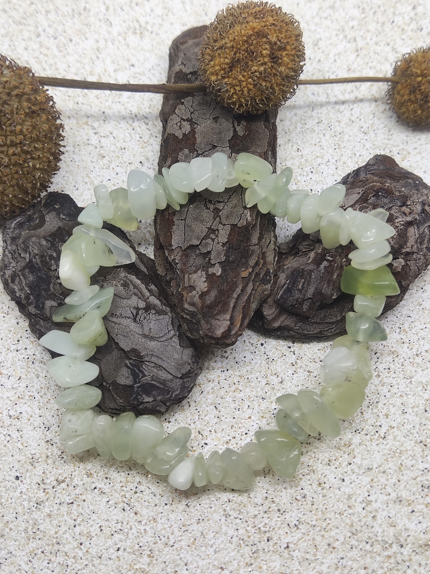 Pulseira de pedras verdes irregulares sobre areia com casca de árvore castanha e plantas secas.