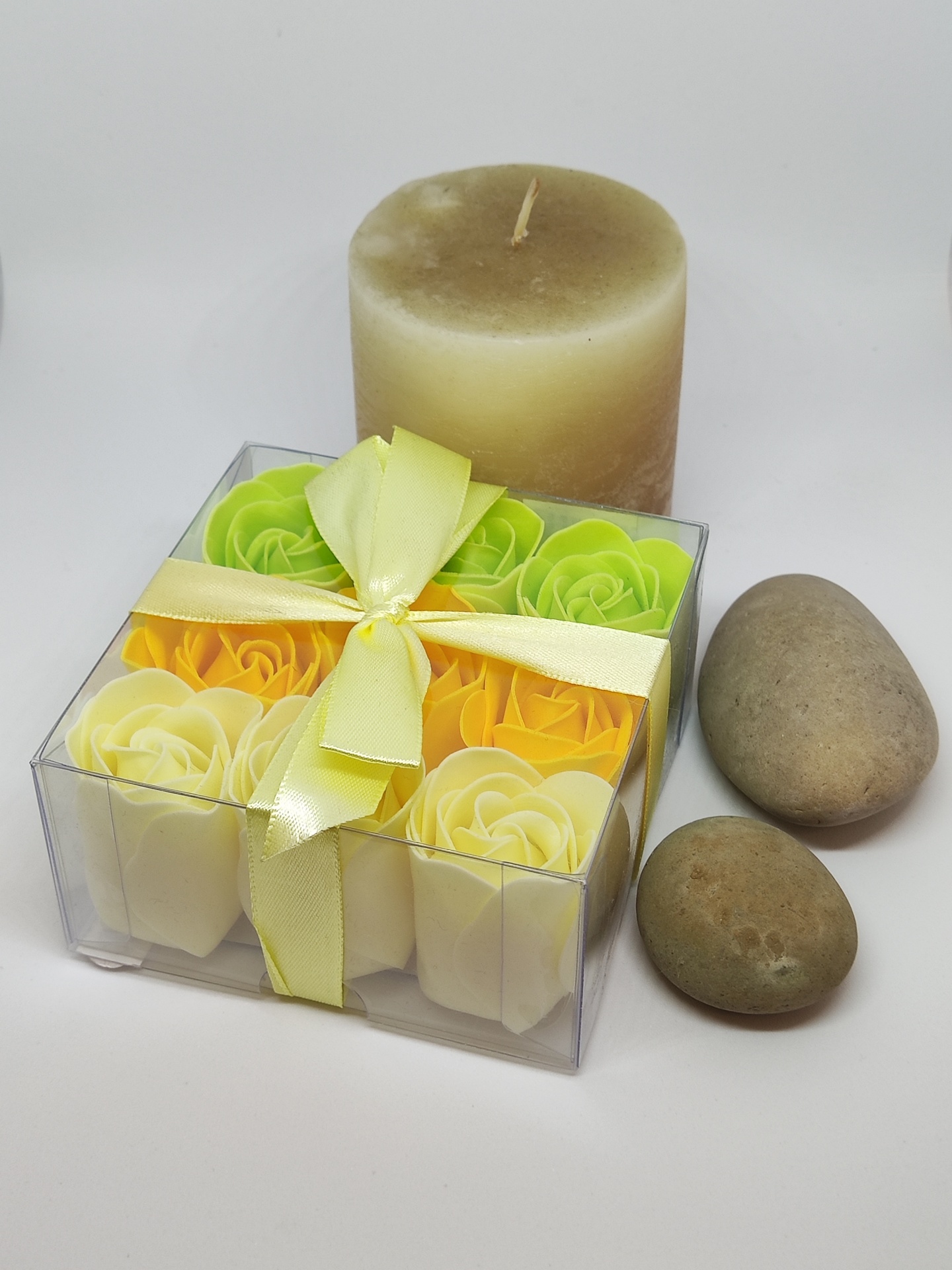Caixa com flores decorativas coloridas, vela bege cilíndrica e duas pedras ovais em fundo branco