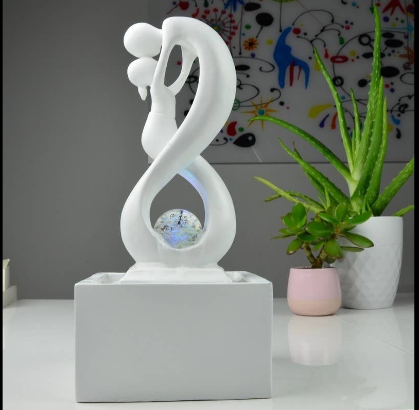 Escultura abstrata branca de casal com esfera decorativa no centro, sobre base branca, com plantas e pintura ao fundo