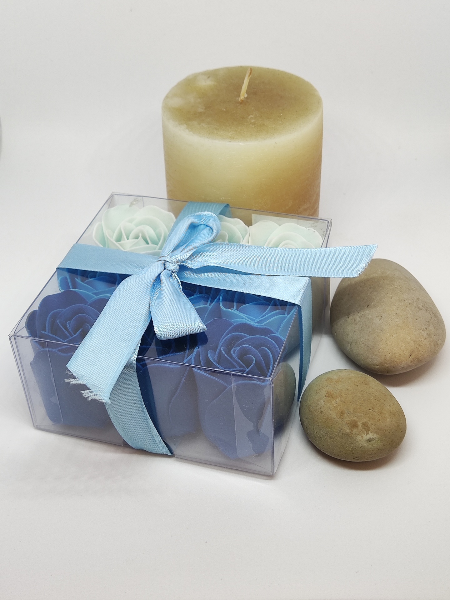Vela castanha clara, pedras naturais e caixa transparente com flores de sabão azuis e brancas amarrada com fita azul