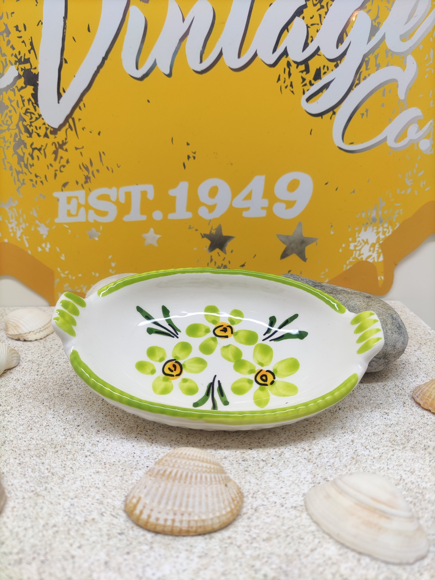 Prato oval de cerâmica decorado com flores verdes, sobre mesa areia com conchas e fundo amarelo com texto