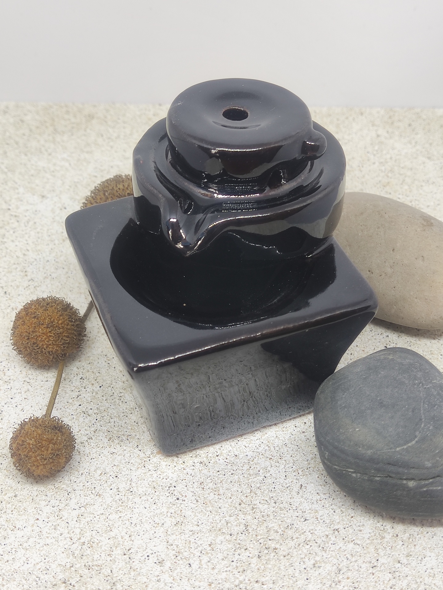 Frasco de vidro preto quadrado com tampa redonda junto a pedras e frutos secos.