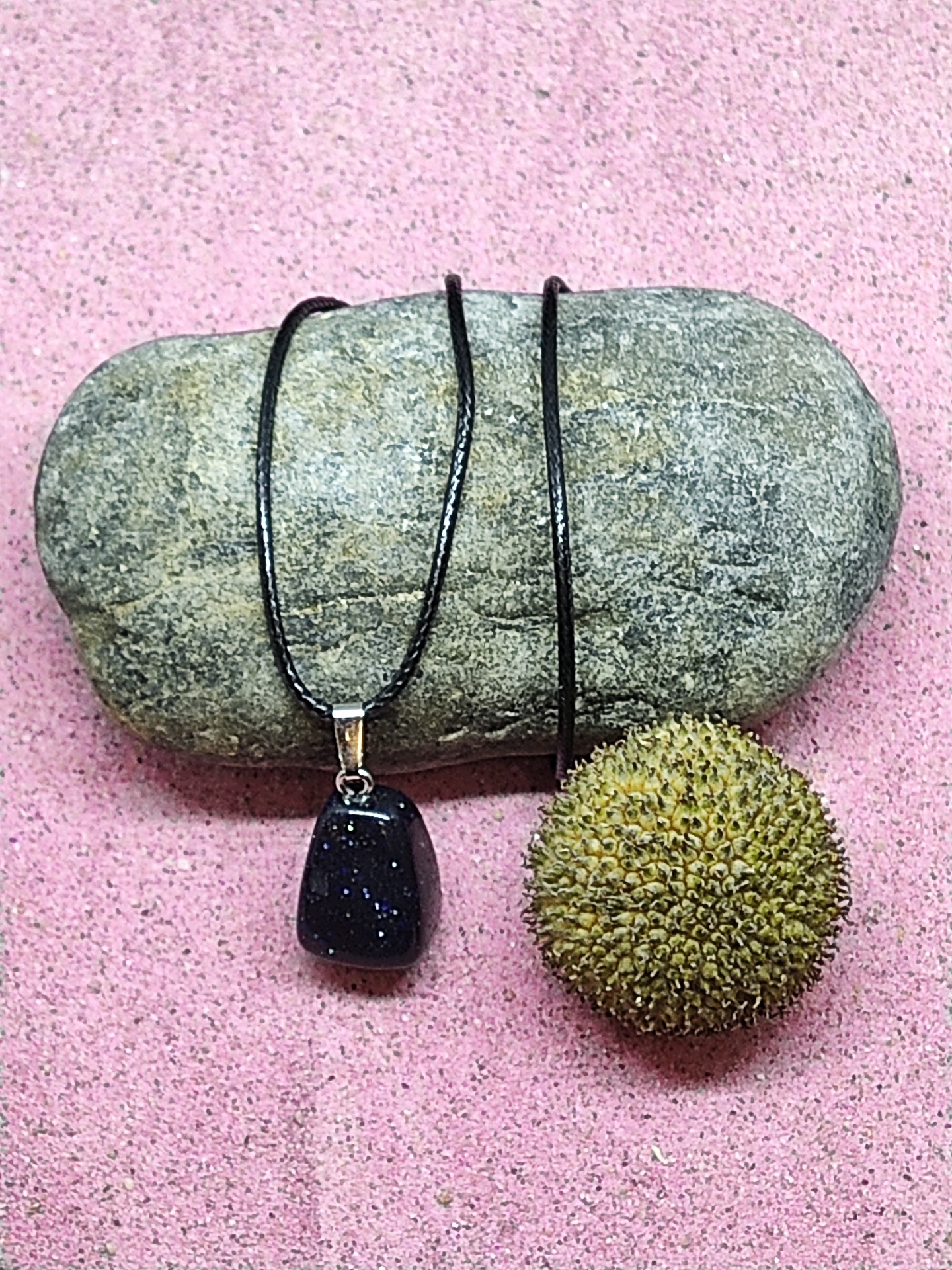 Colar com pendente de pedra preta sobre pedra cinzenta e fruto espinhento em fundo rosa.