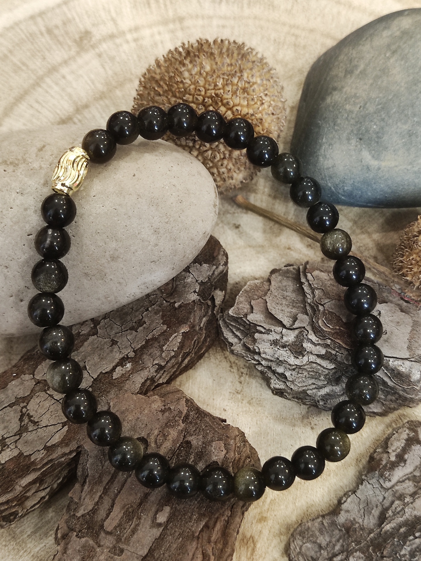 Pulseira de contas pretas com detalhe dourado sobre troncos, pedras e frutos secos