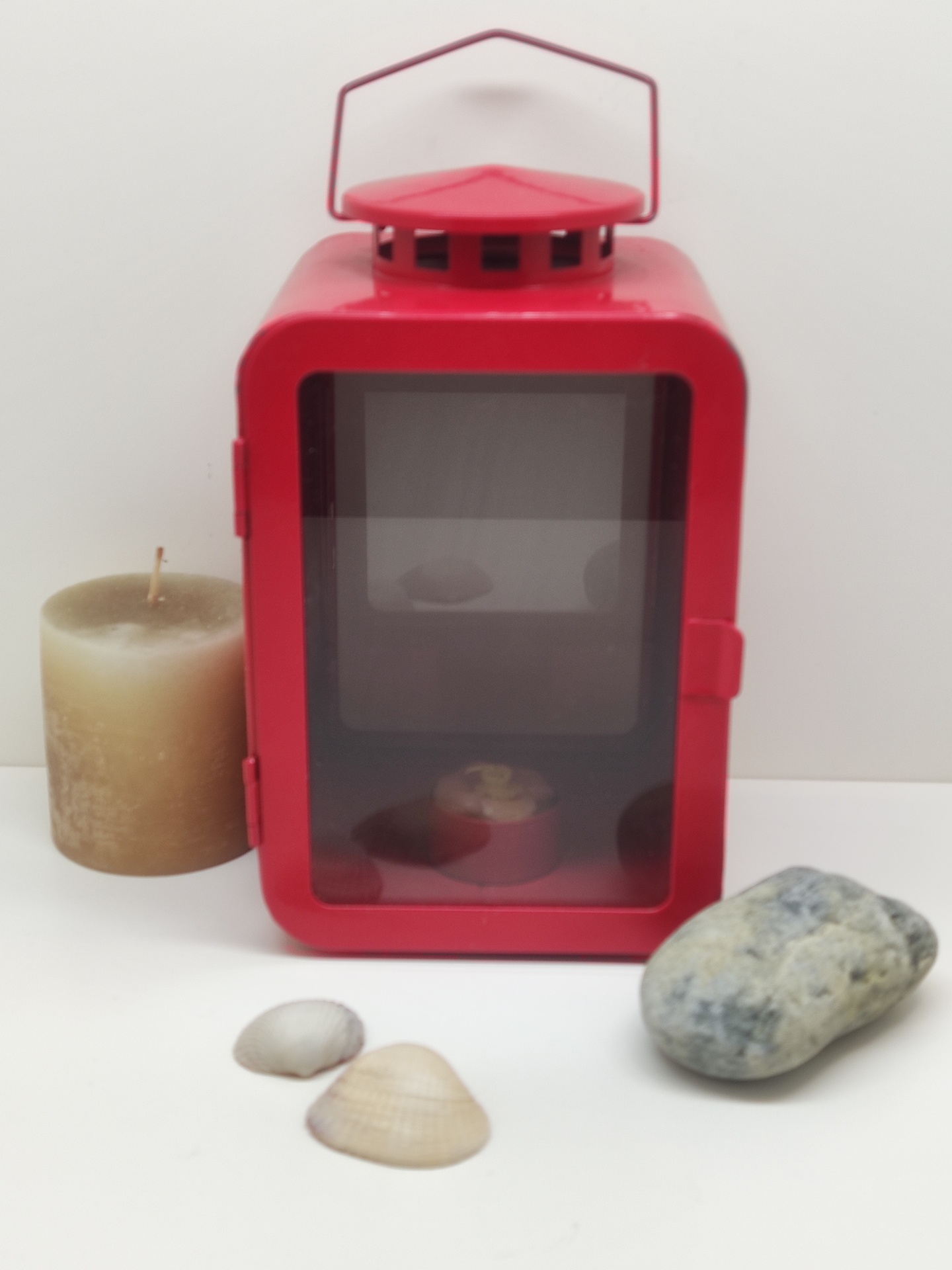 Lanterna metálica vermelha com vela castanha, conchas e pedra sobre fundo branco