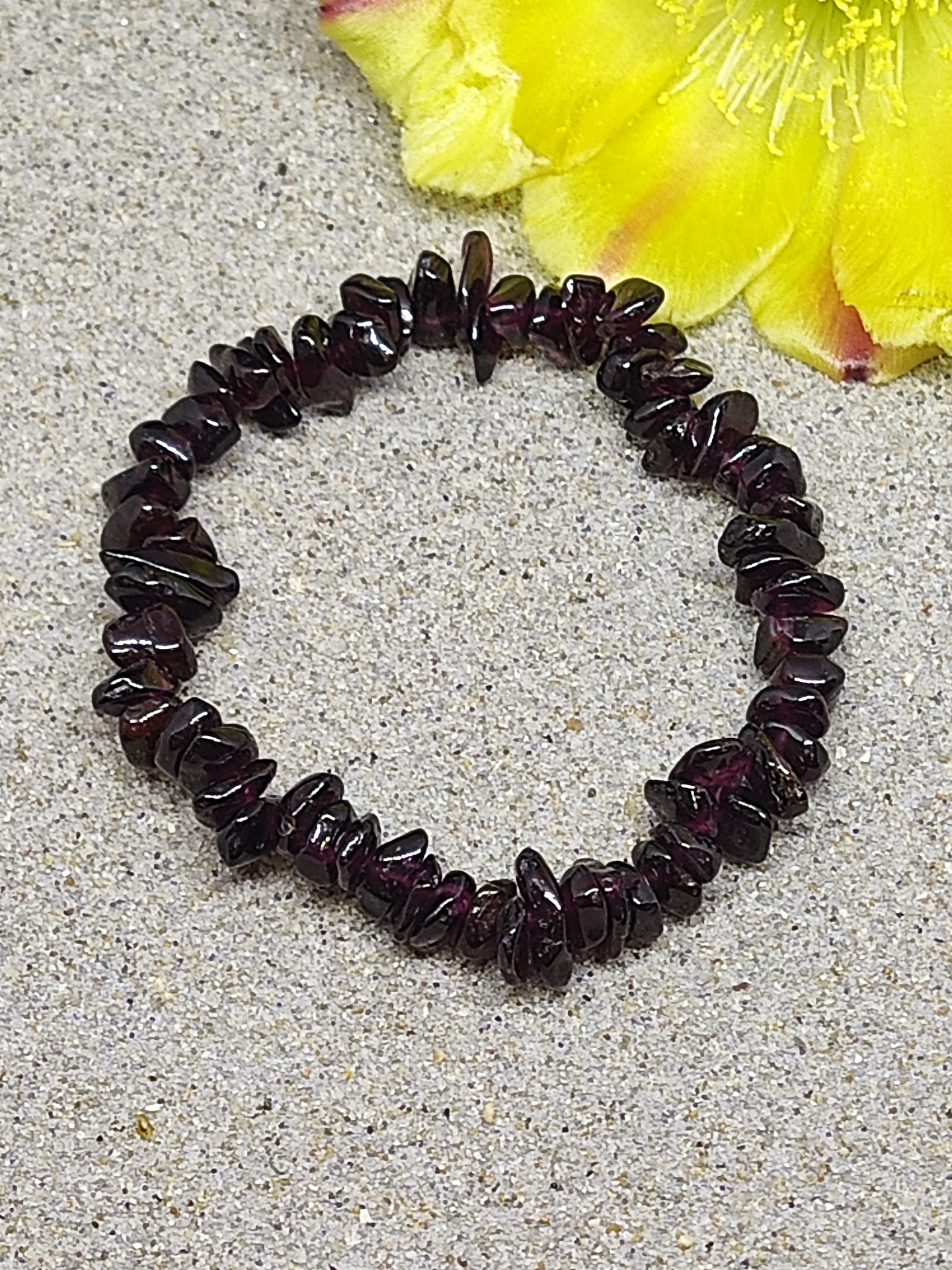 Pulseira de pedras grená escuras sobre superfície cinza com flor amarela ao lado