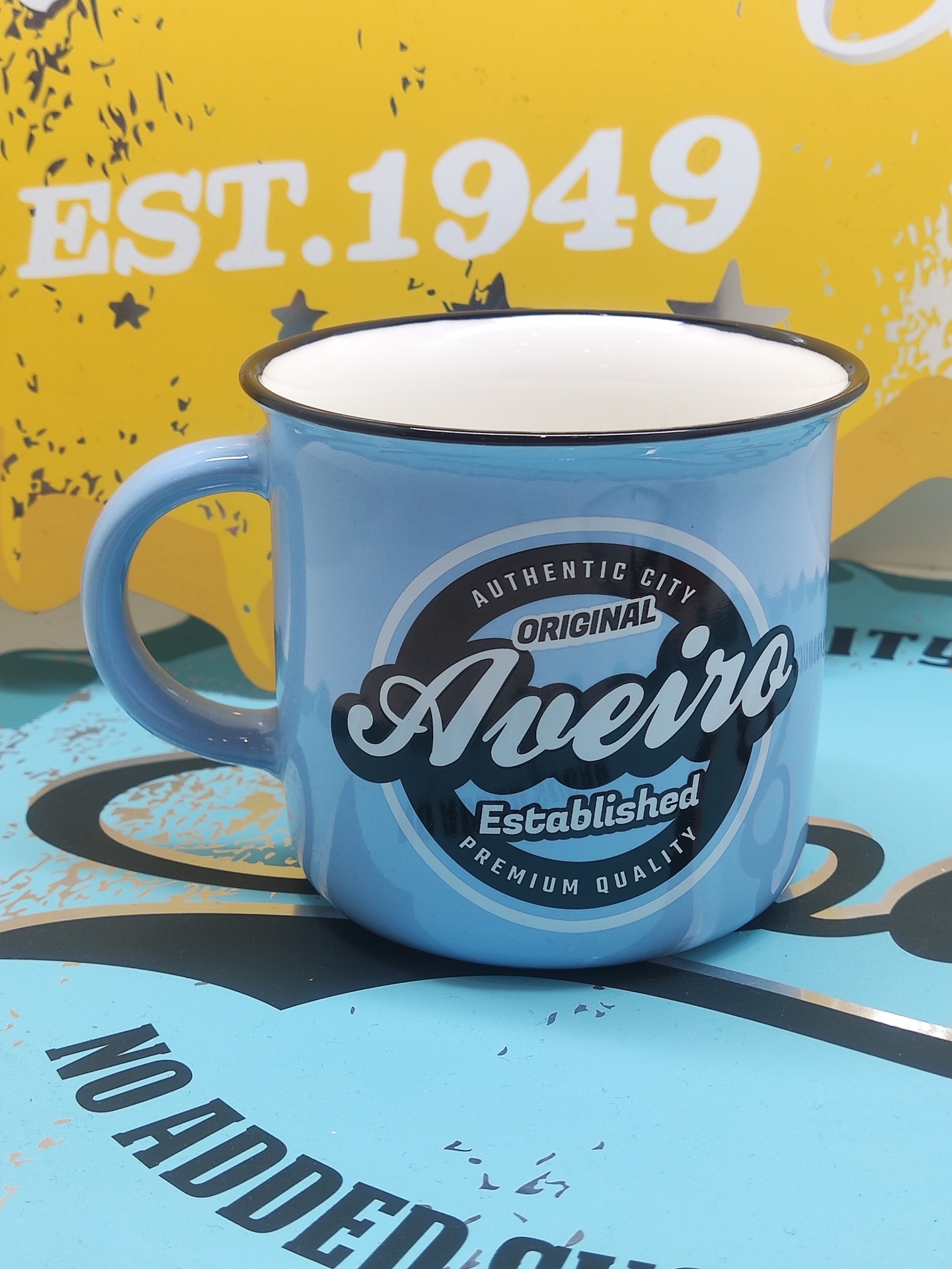 Caneca azul clara em metal esmaltado com texto Original Aveiro em fundo amarelo e azul