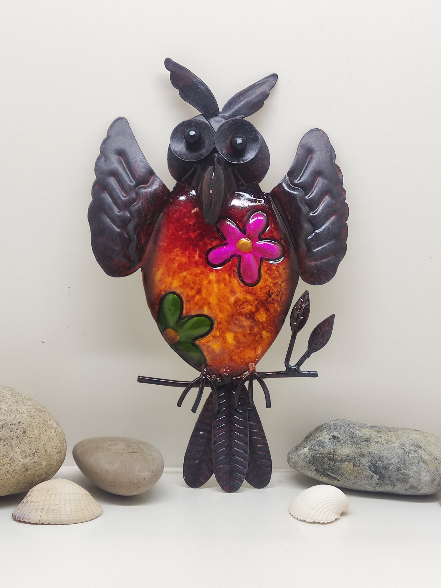 Escultura colorida de coruja em metal decorada com flores, sobre superfície com pedras e conchas.
