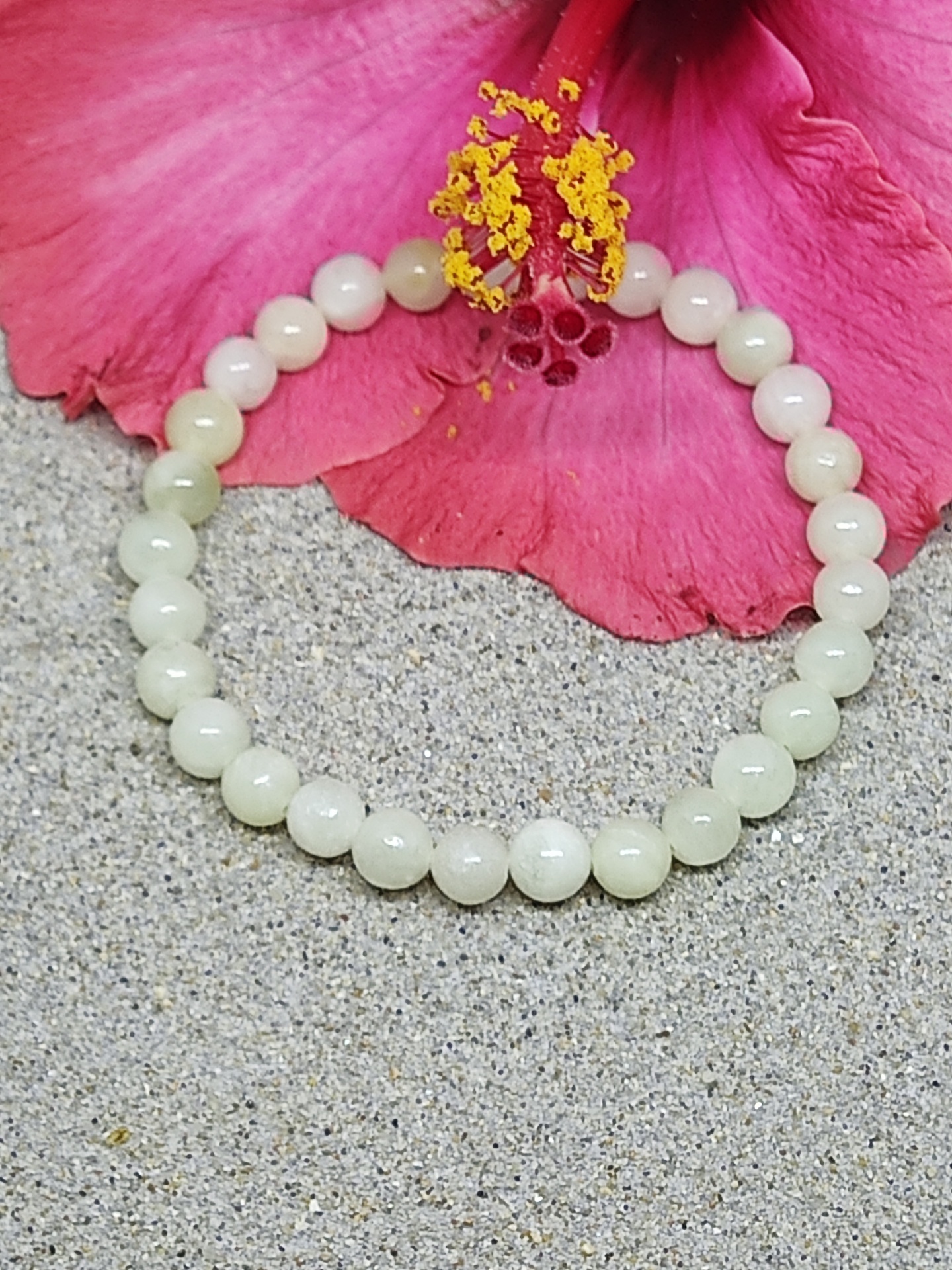 Pulseira branca de contas numa areia com flor rosa hibisco ao fundo