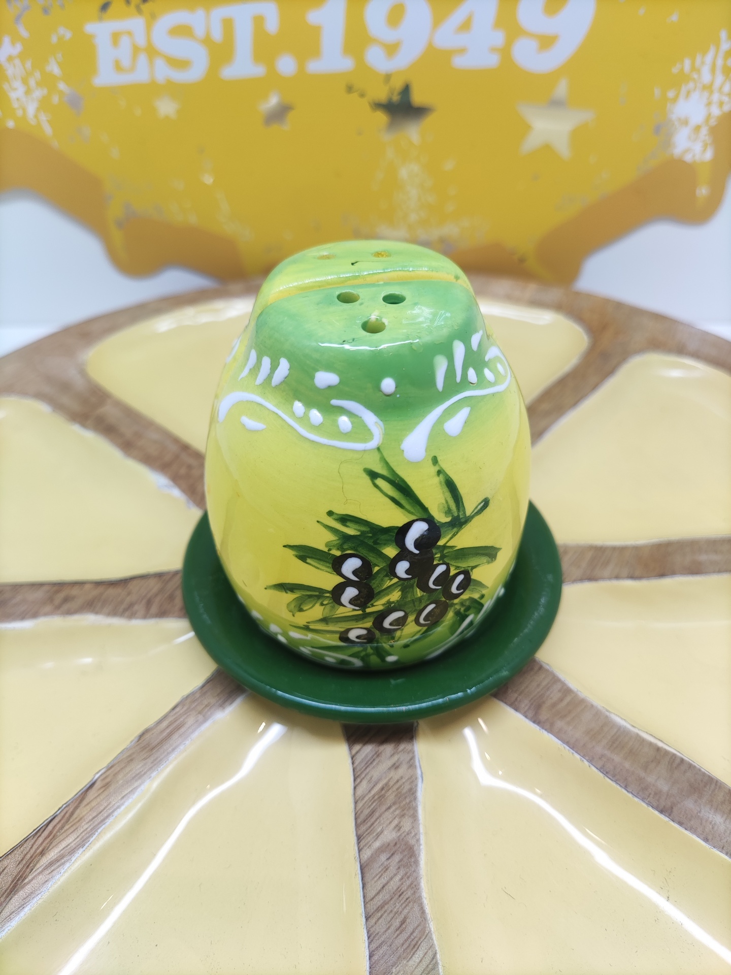 Saleiro cerâmico verde e amarelo com decoração de pinhas, sobre prato com padrão amarelo e castanho, com letreiro amarelo ao fundo