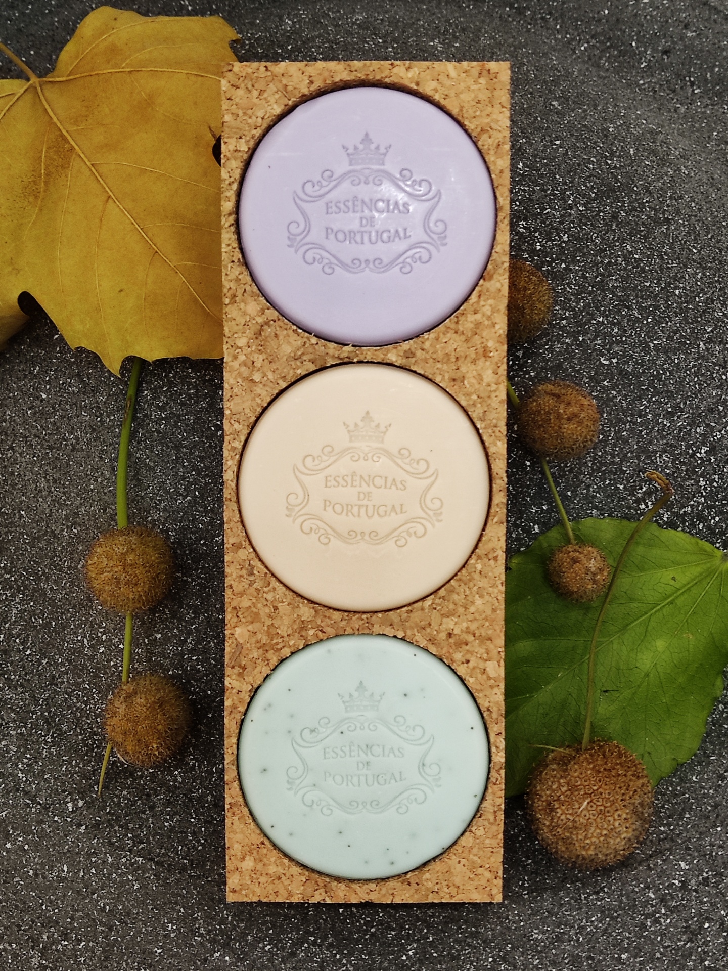 Três sabonetes coloridos em suporte de cortiça com folhas e bolotas ao redor