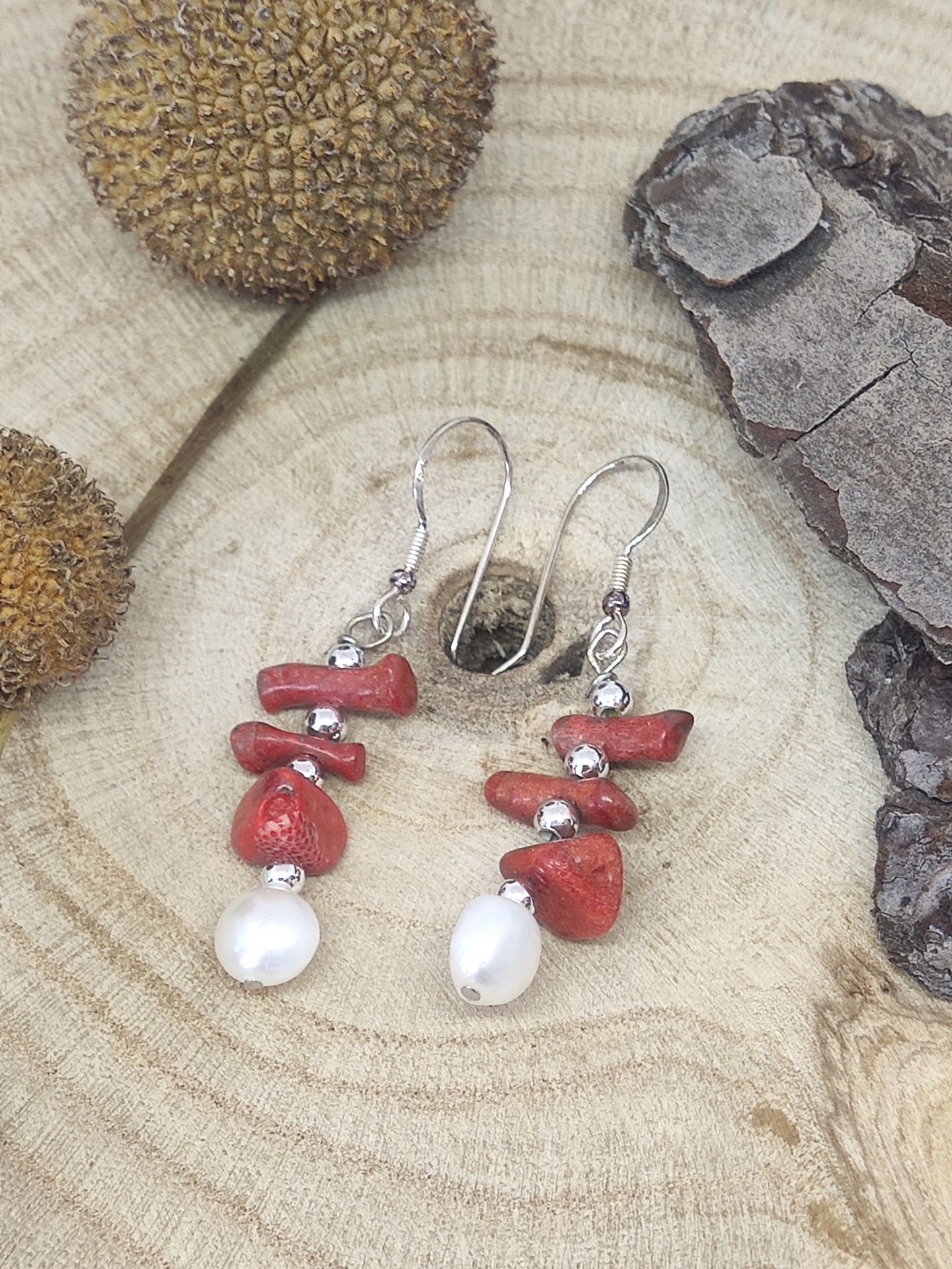 Brincos com pedras vermelhas, contas prateadas e pérolas brancas sobre madeira e elementos naturais.