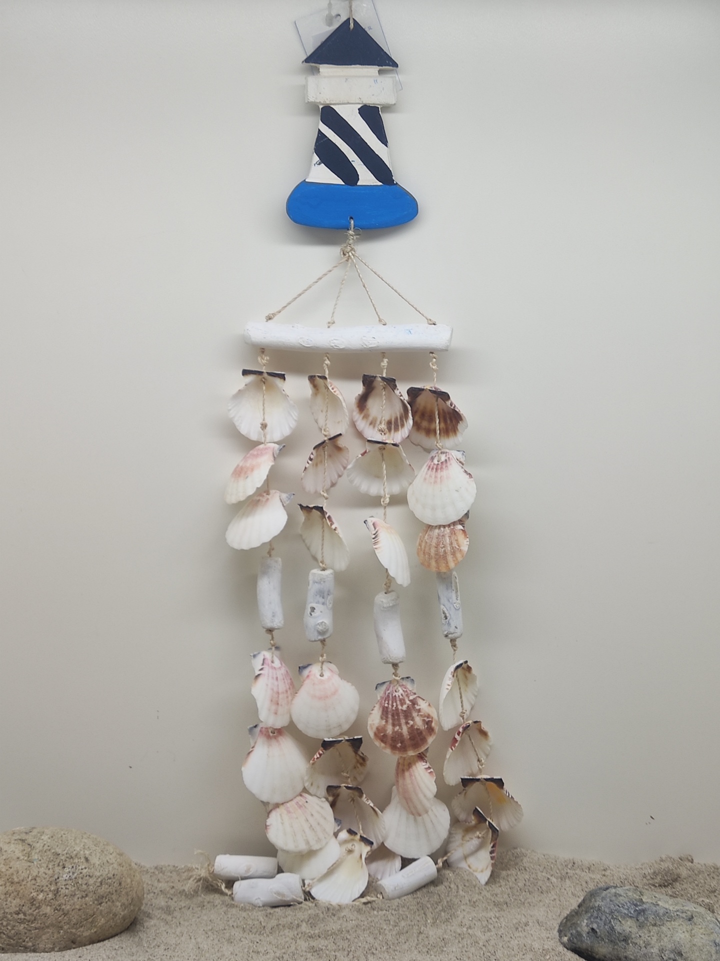 Suspensão decorativa com conchas, madeira pintada de azul e branco, pendurada numa parede clara sobre areia e pedras