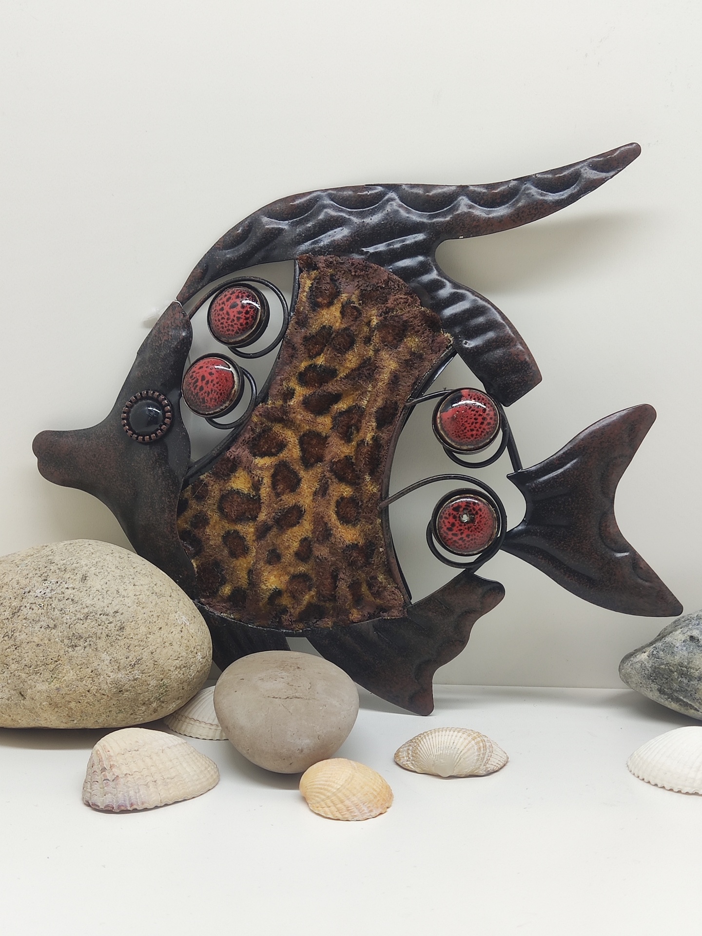 Escultura de peixe em metal e tecido com padrão de pele de leopardo sobre superfície branca com conchas e pedras