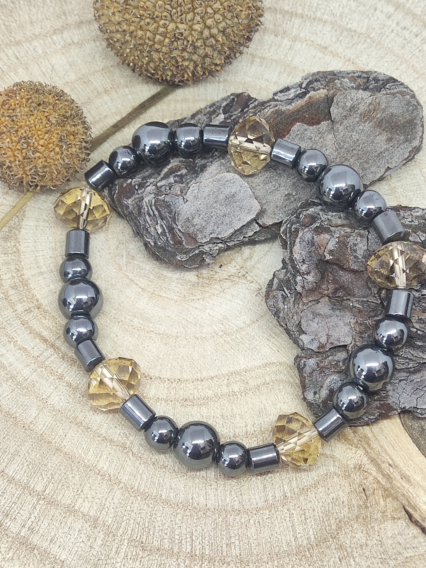 Pulseira com contas metálicas cinza escuro e âmbar sobre fundo de madeira com cascas e frutos secos