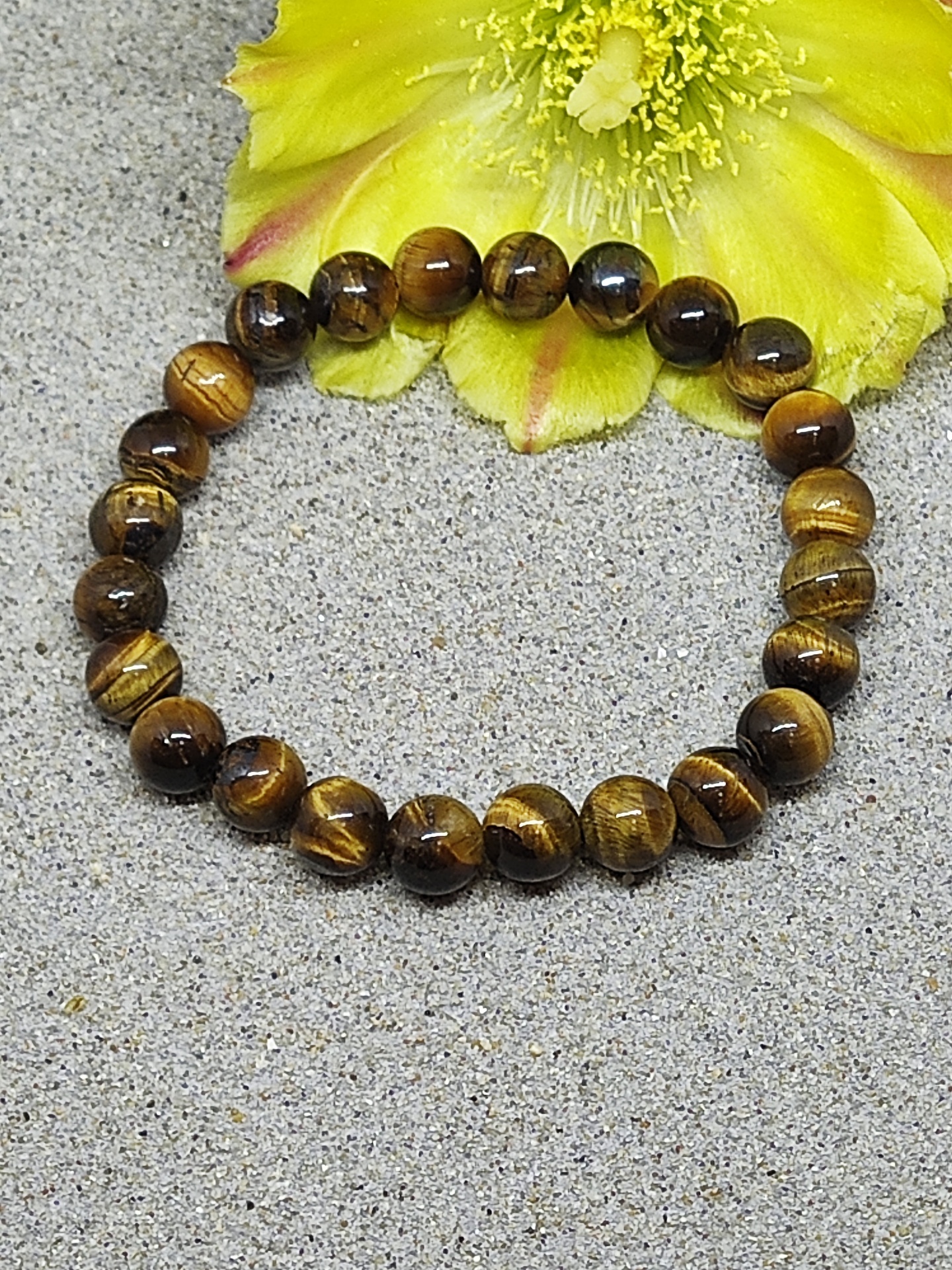 Pulseira de contas de pedra olho de tigre castanha com reflexos dourados sobre superfície cinzenta e flor amarela