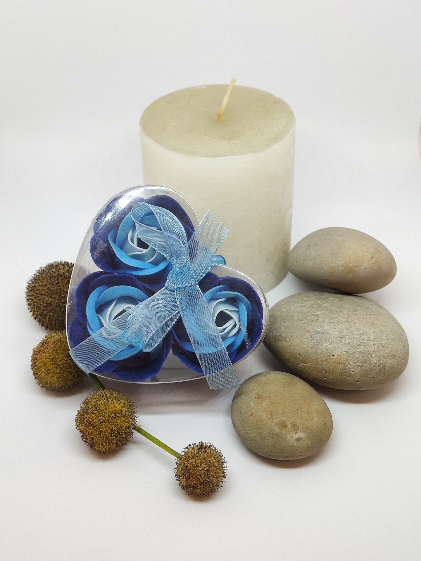 Conjunto decorativo com vela branca, flores de tecido azuis e pedras cinzentas sobre fundo branco.