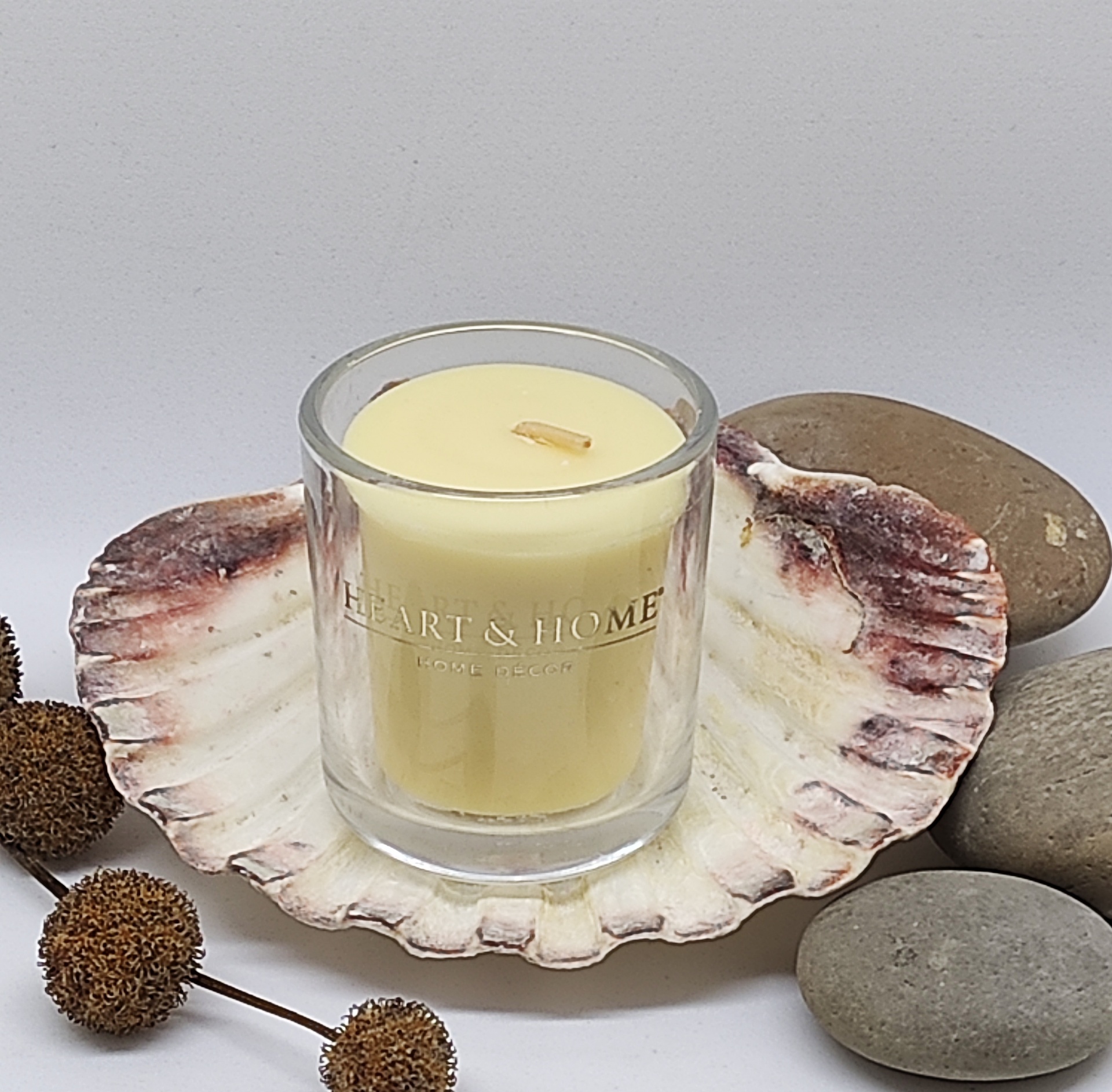 Vela amarela em copo de vidro com inscrição Heart & Home sobre concha grande com pedras e flores secas ao lado