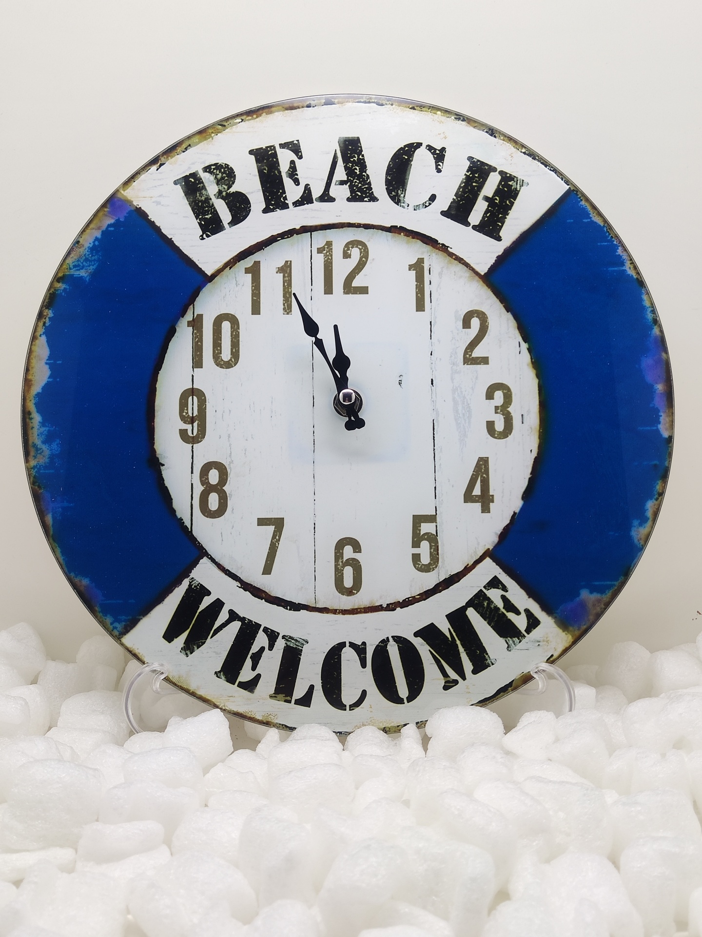 Relógio decorativo redondo tipo bóia salva-vidas com texto Beach Welcome em fundo branco e azul.