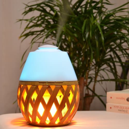 Difusor aromático azul e madeira com luz laranja numa mesa branca, planta verde ao fundo