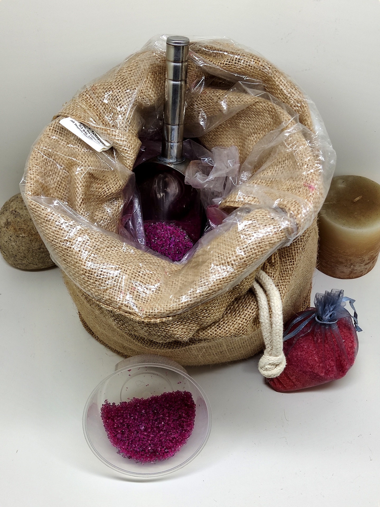 Saco de ráfia com frasco de líquido, granulado rosa, colher, velas e saco vermelho sobre superfície branca