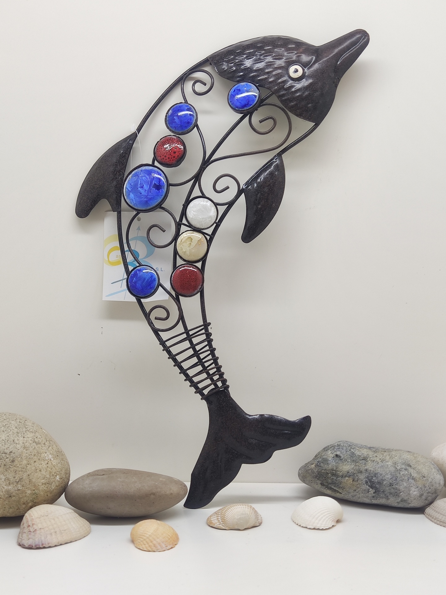 Escultura de golfinho em metal com detalhes coloridos colocada sobre uma superfície branca com pedras e conchas.