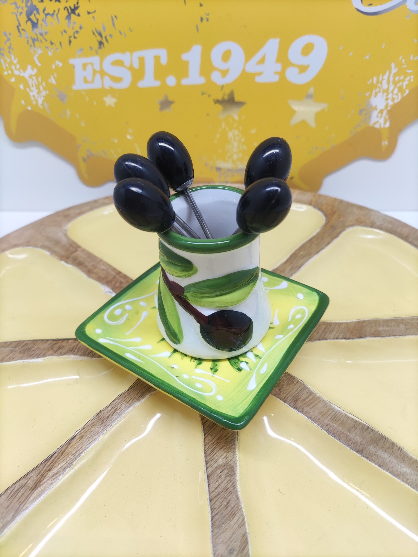 Conjunto de jarro e prato cerâmicos com espetos metálicos com cabos pretos, sobre prato redondo dividido, fundo com placa amarela EST.1949