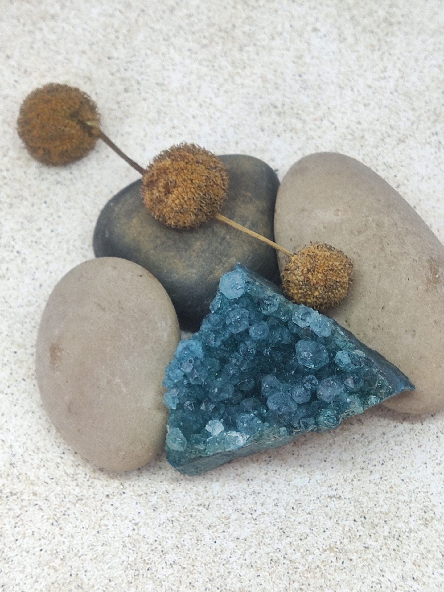 Pedra mineral azul com cristais sobre pedras lisas e flores secas em superfície arenosa.