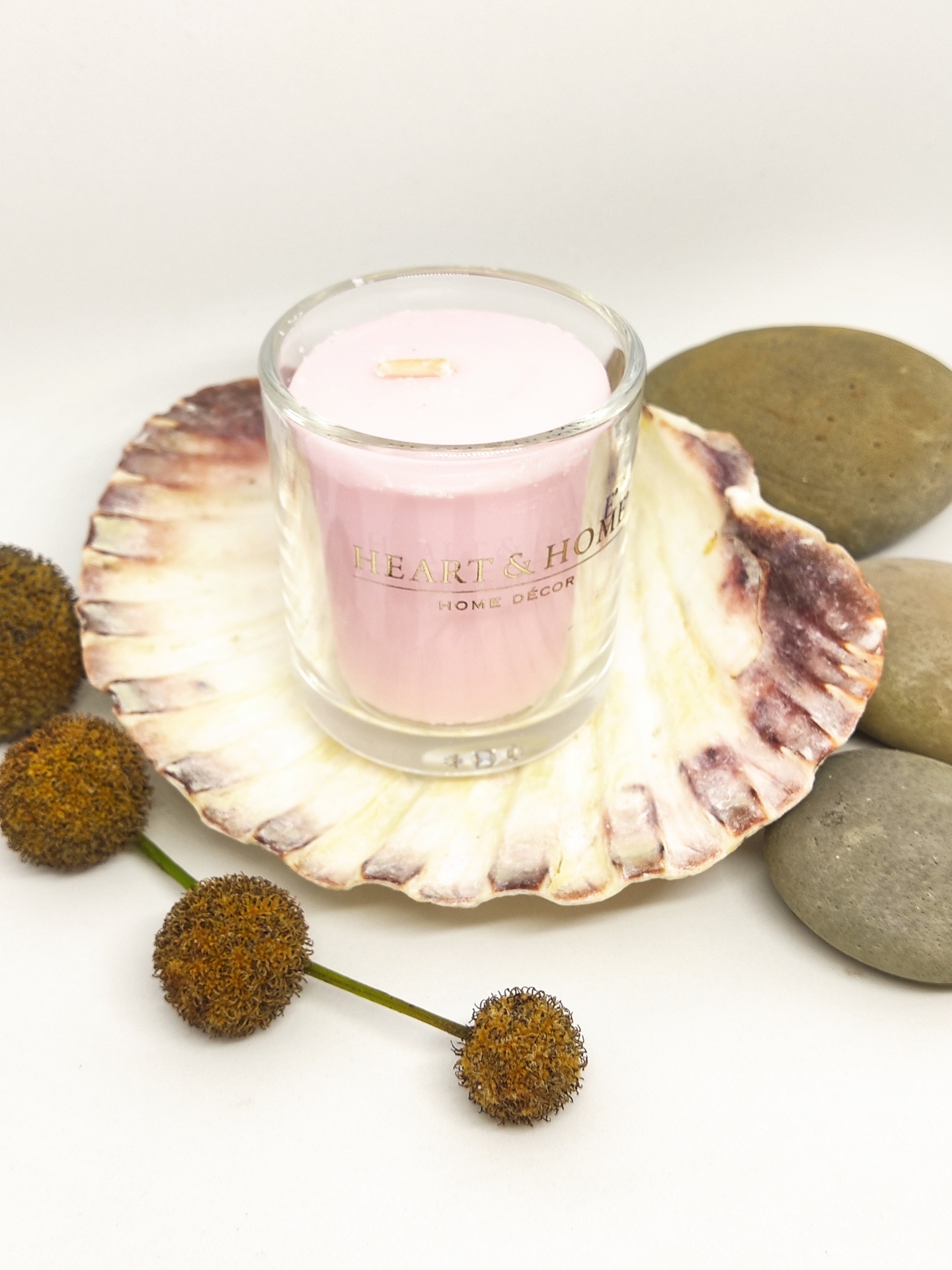 Vela rosa em copo de vidro sobre concha grande com pedras e flores secas