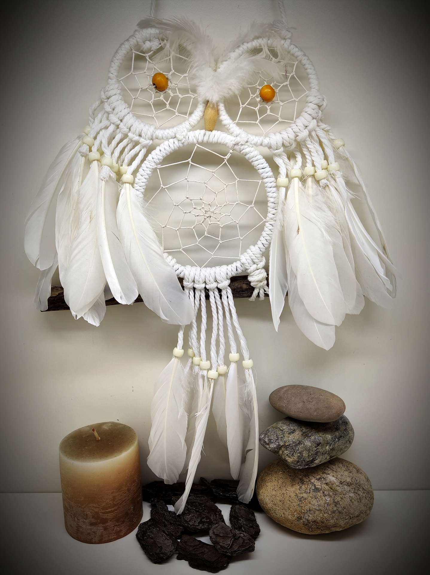 Pendurado decorativo em forma de coruja com penas brancas e miçangas amarelas, vela e pedras ao lado.