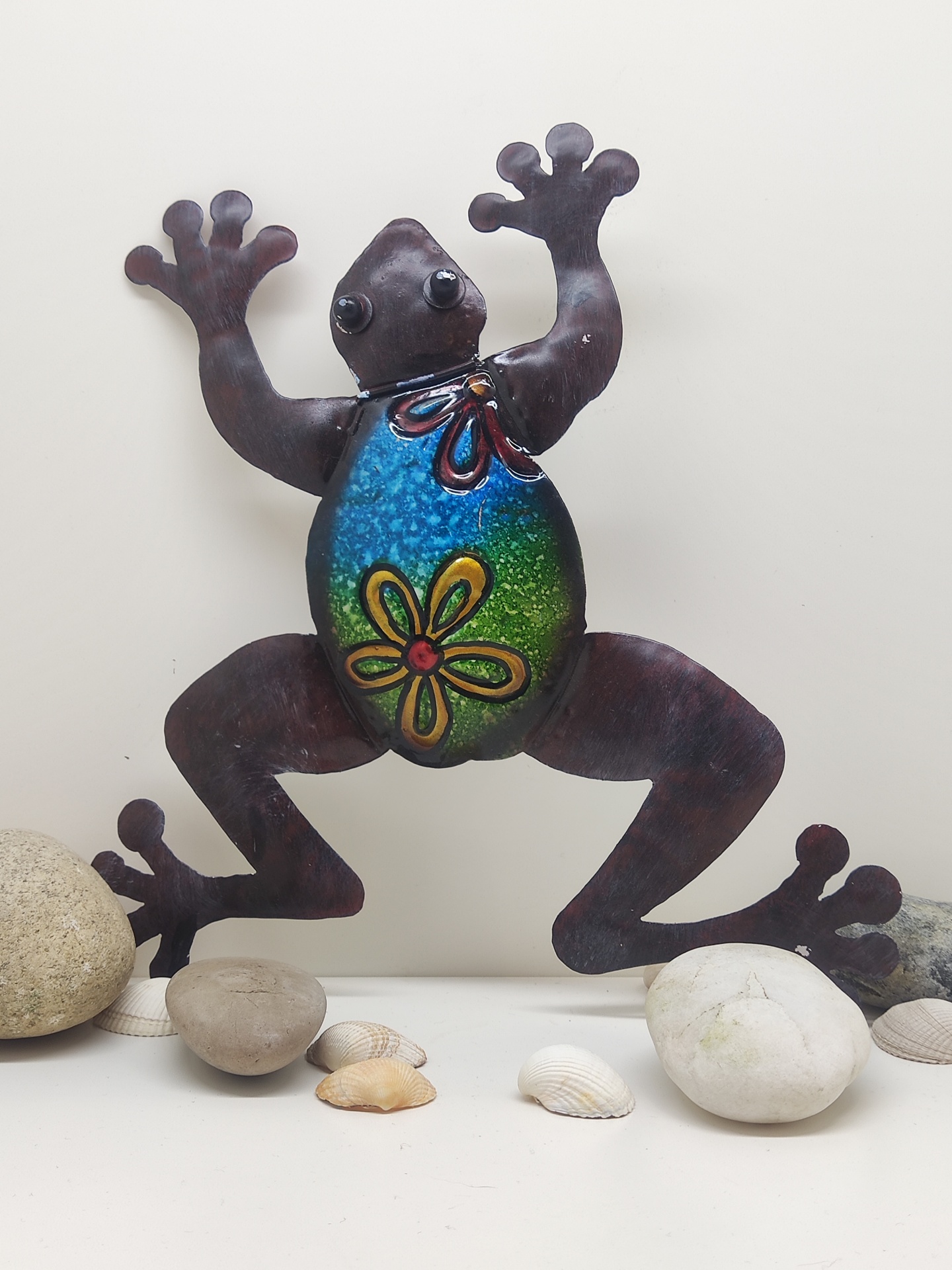 Escultura metálica colorida de rã decorativa com fundo branco e pedras ao redor