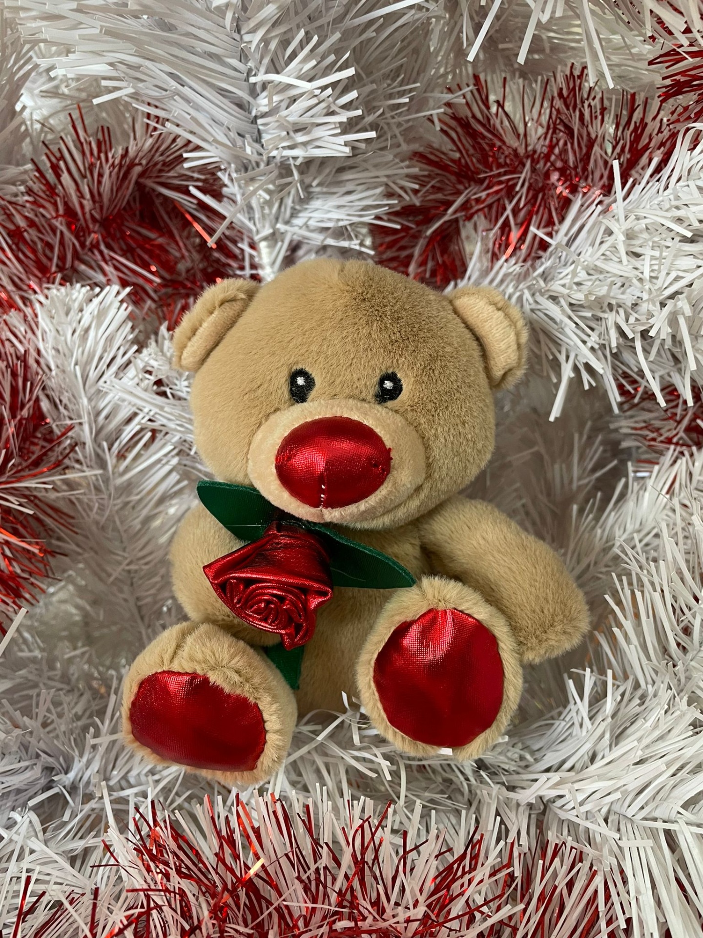 Ursinho de peluche castanho com detalhes vermelhos segurando uma rosa entre decoração natalícia branca e vermelha