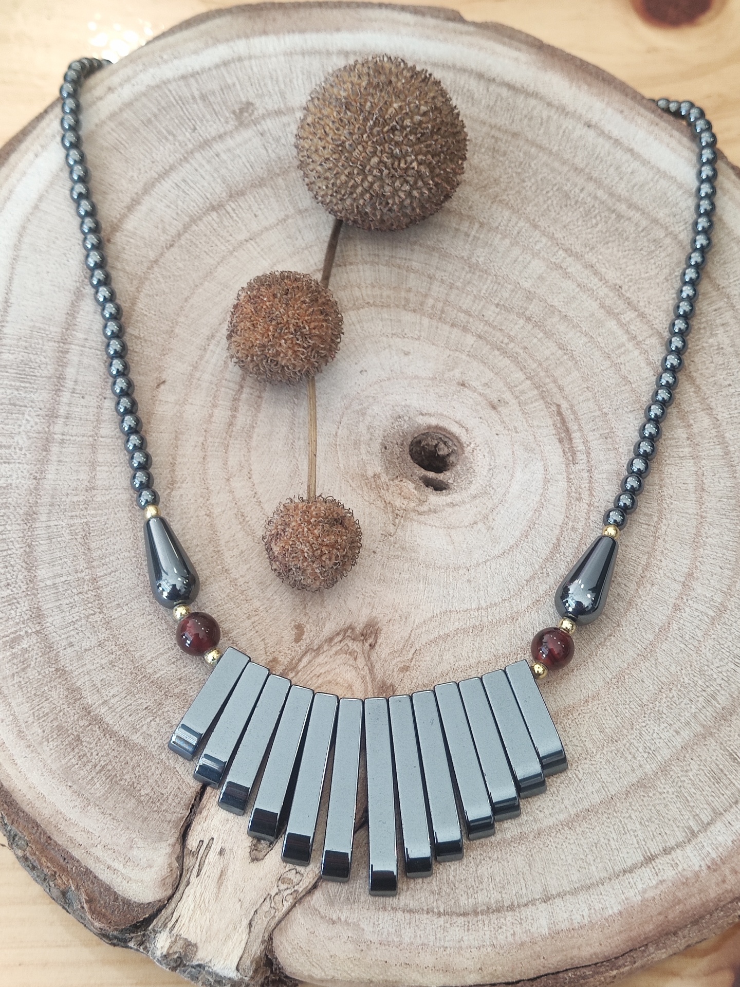 Colar em contas escuras com pendentes cinza metálico sobre base de madeira clara com frutos secos.