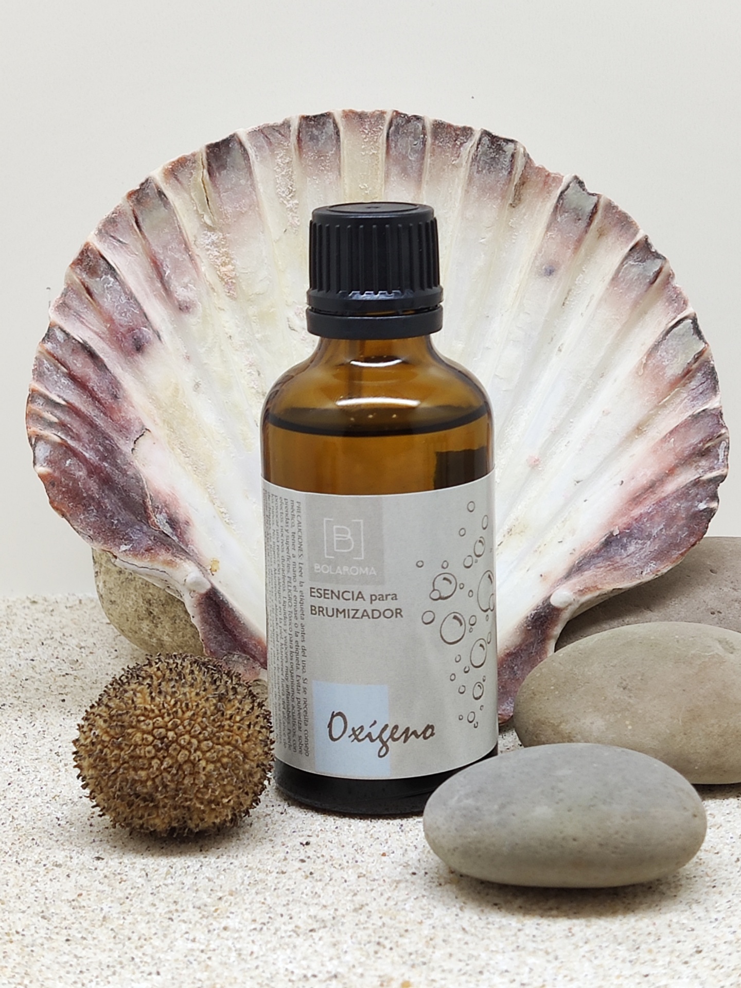 Frasco de essência para brumizador em vidro âmbar com tampa preta, sobre areia, com concha e pedras ao fundo.