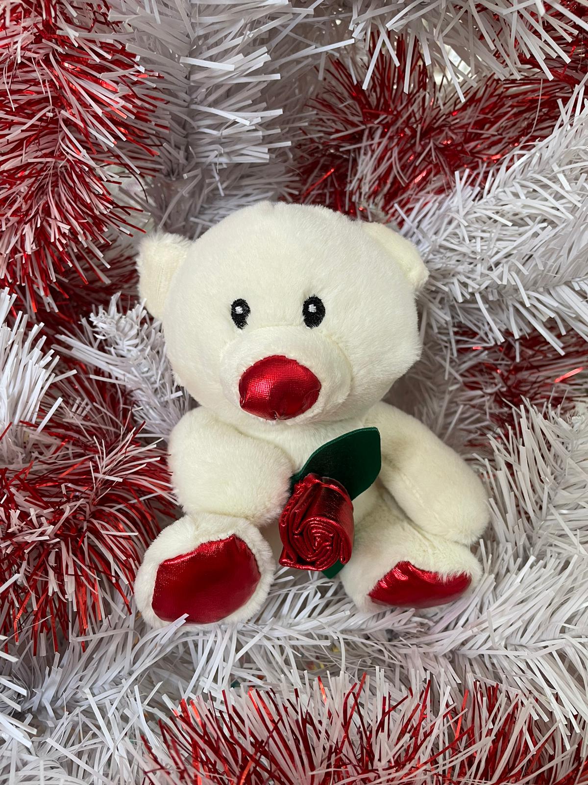 Ursinho de peluche branco com nariz e patas vermelhas segurando rosa vermelha sobre decoração de ramos brancos e vermelhos.