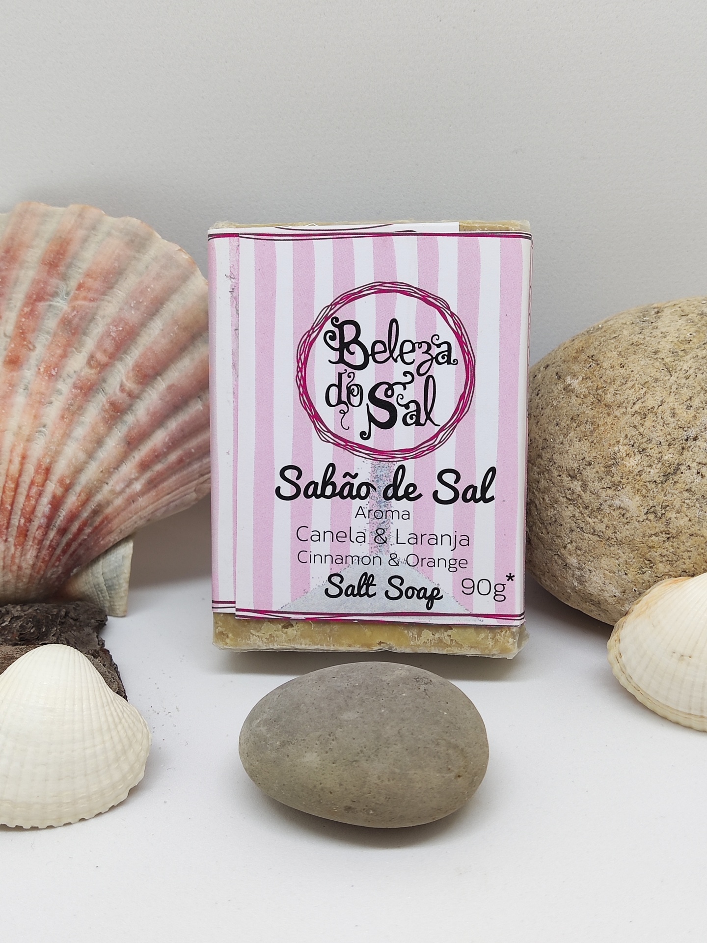 Sabão de sal embalado com rótulo rosa e branco listado entre pedras e conchas