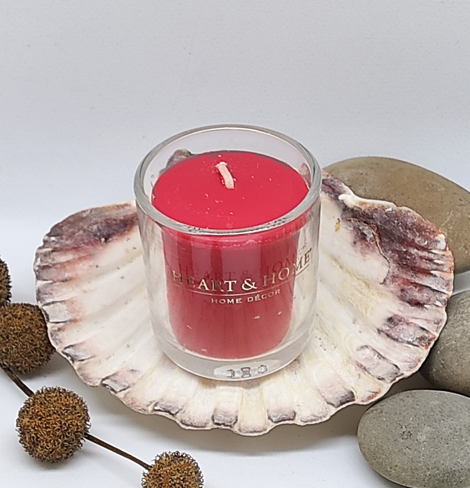 Vela vermelha em copo de vidro com texto HEART & HOME, numa concha decorativa com pedras e elementos naturais