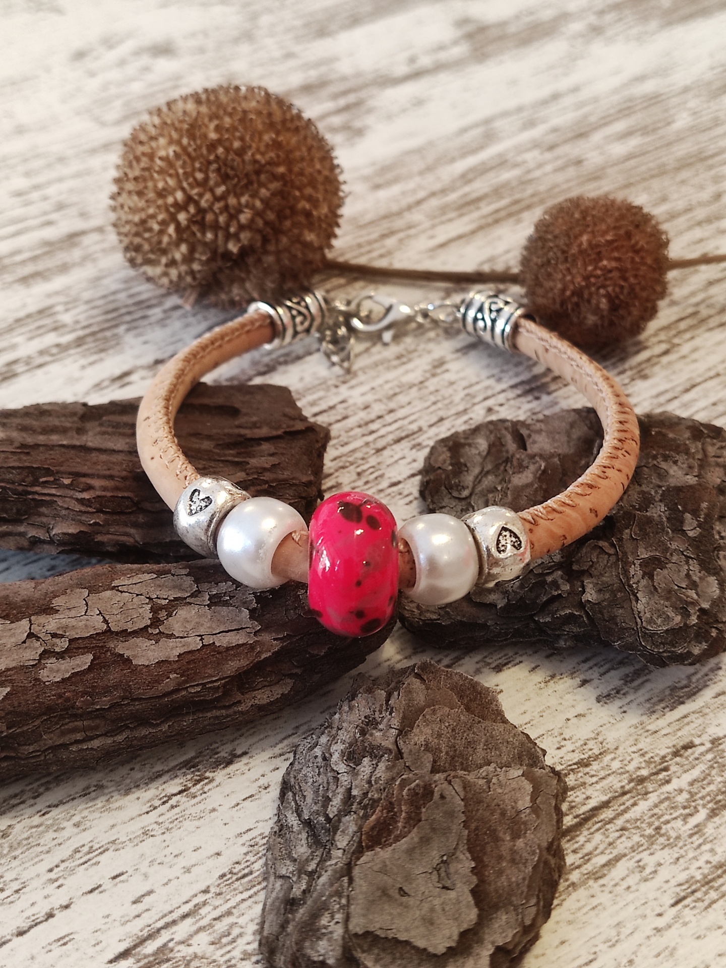 Pulseira de cortiça com contas decorativas e detalhes metálicos prateados, sobre casca de árvore em madeira clara.